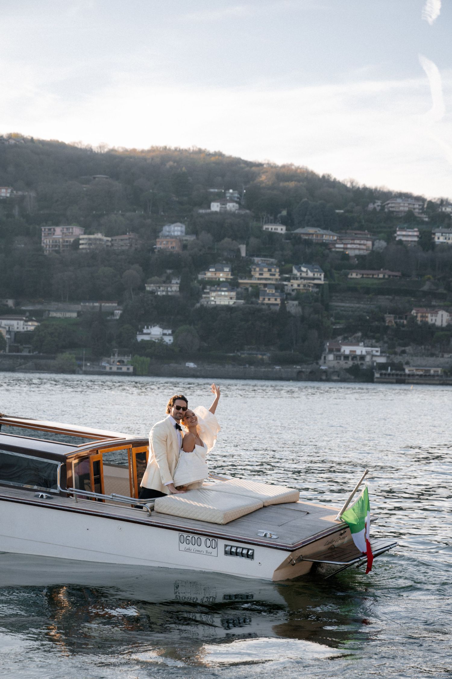 Bride in white dress posing on a luxury wooden boat on Lake Como, Italy, with hillside villas in background.