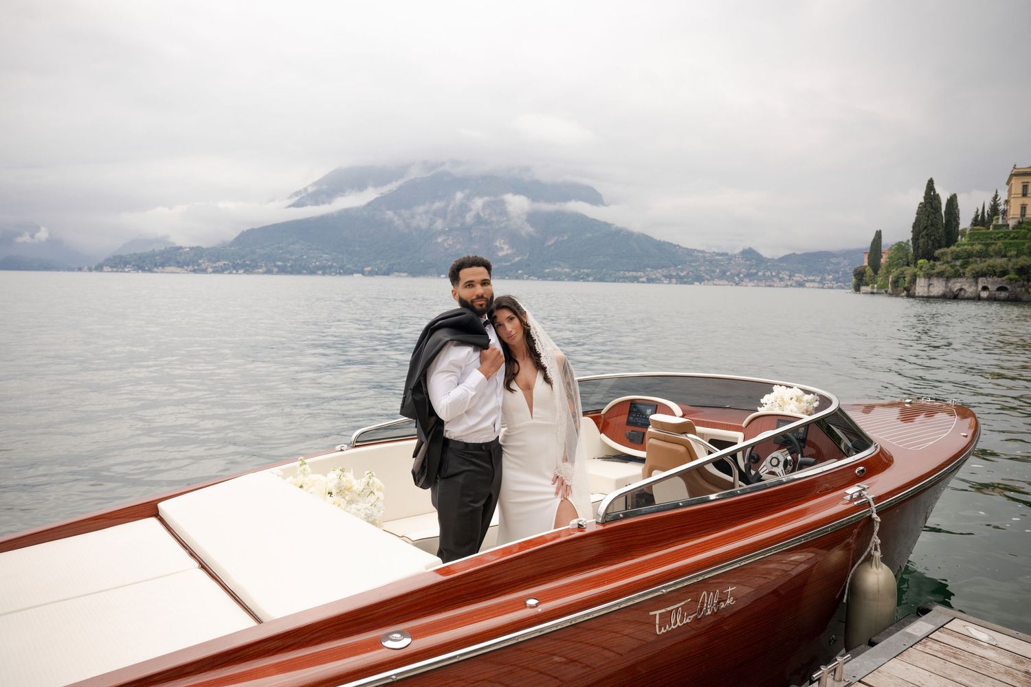Couple posing on a classic wooden boat on Lake Como, Italy, with misty mountains in the background.