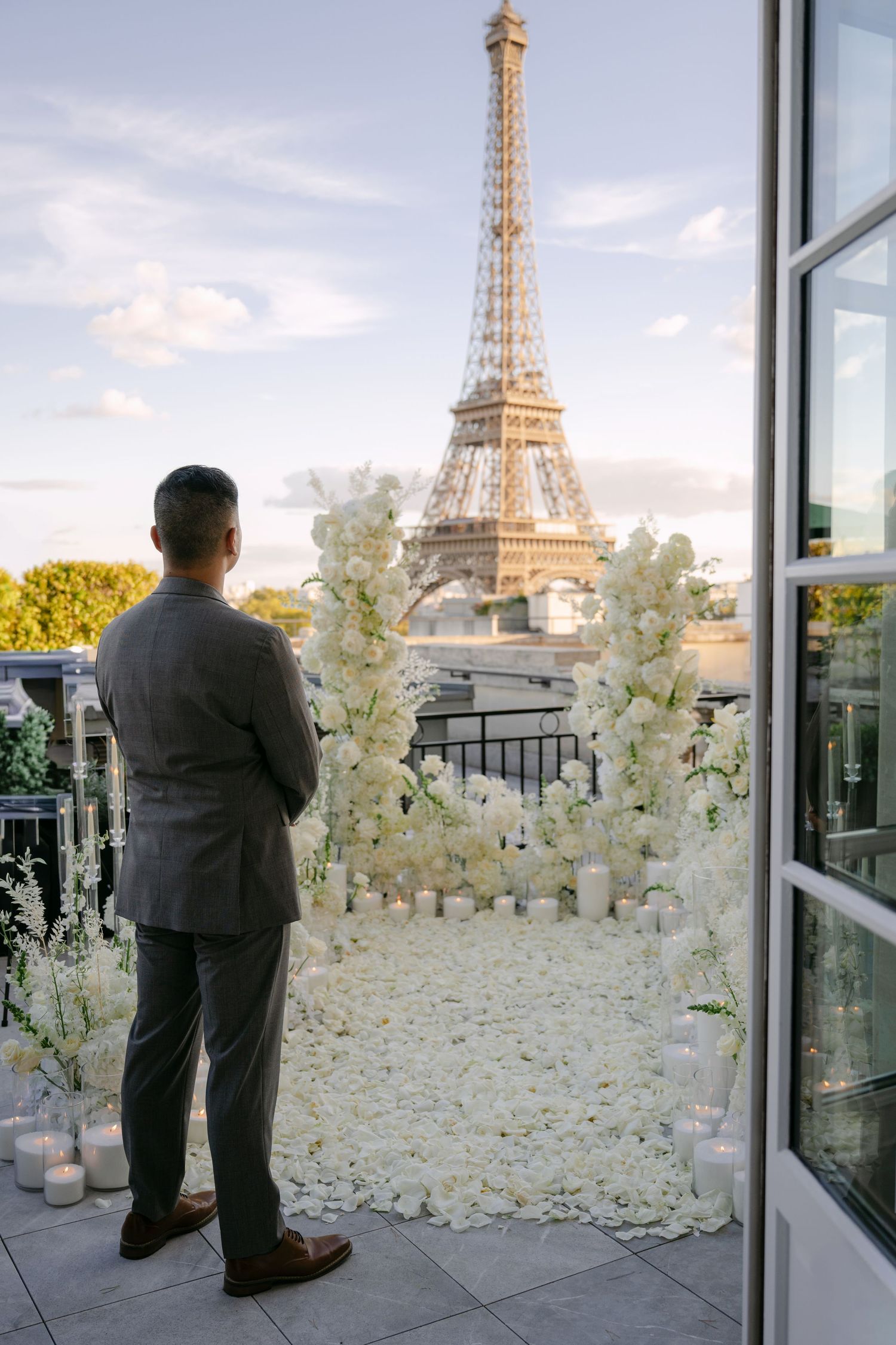 Man in a gray suit stands before a romantic white floral arch and candles, gazing at the Eiffel Tower in Paris.
