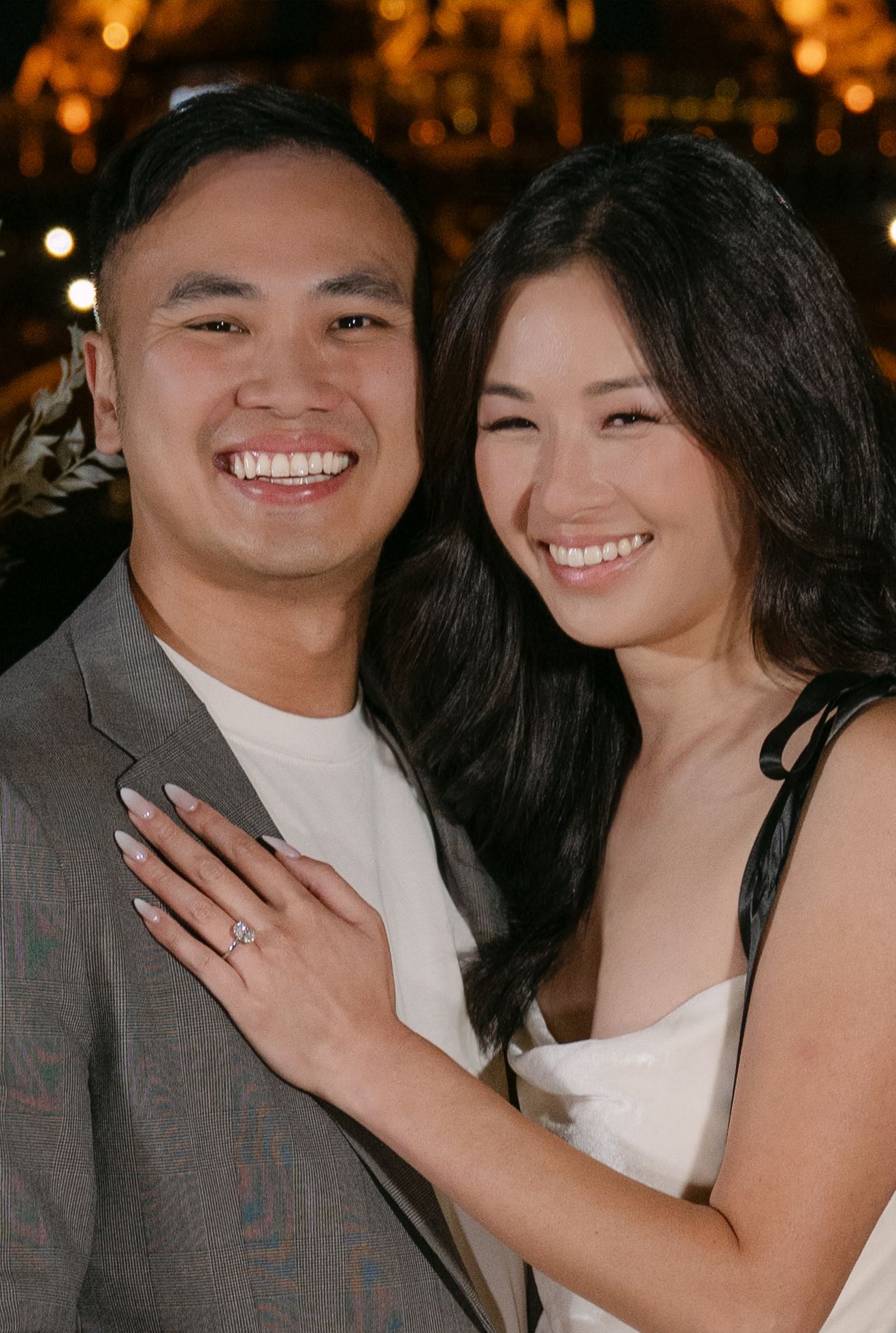A smiling couple embraces closely as the woman shows off a sparkling diamond engagement ring, with warm string lights glowing behind them.