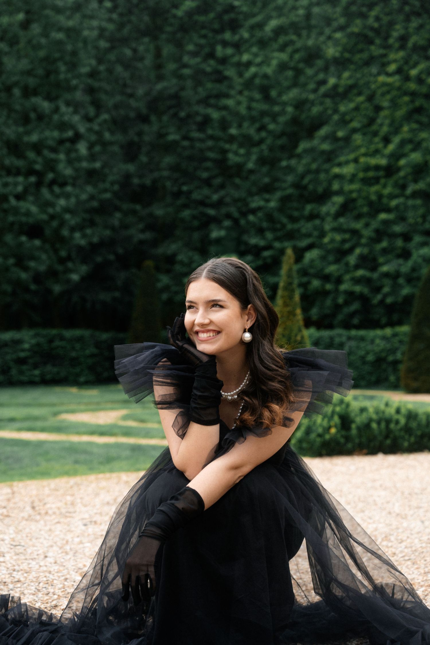 Smiling woman in black tulle off-shoulder gown with gloves poses in formal garden with hedges.