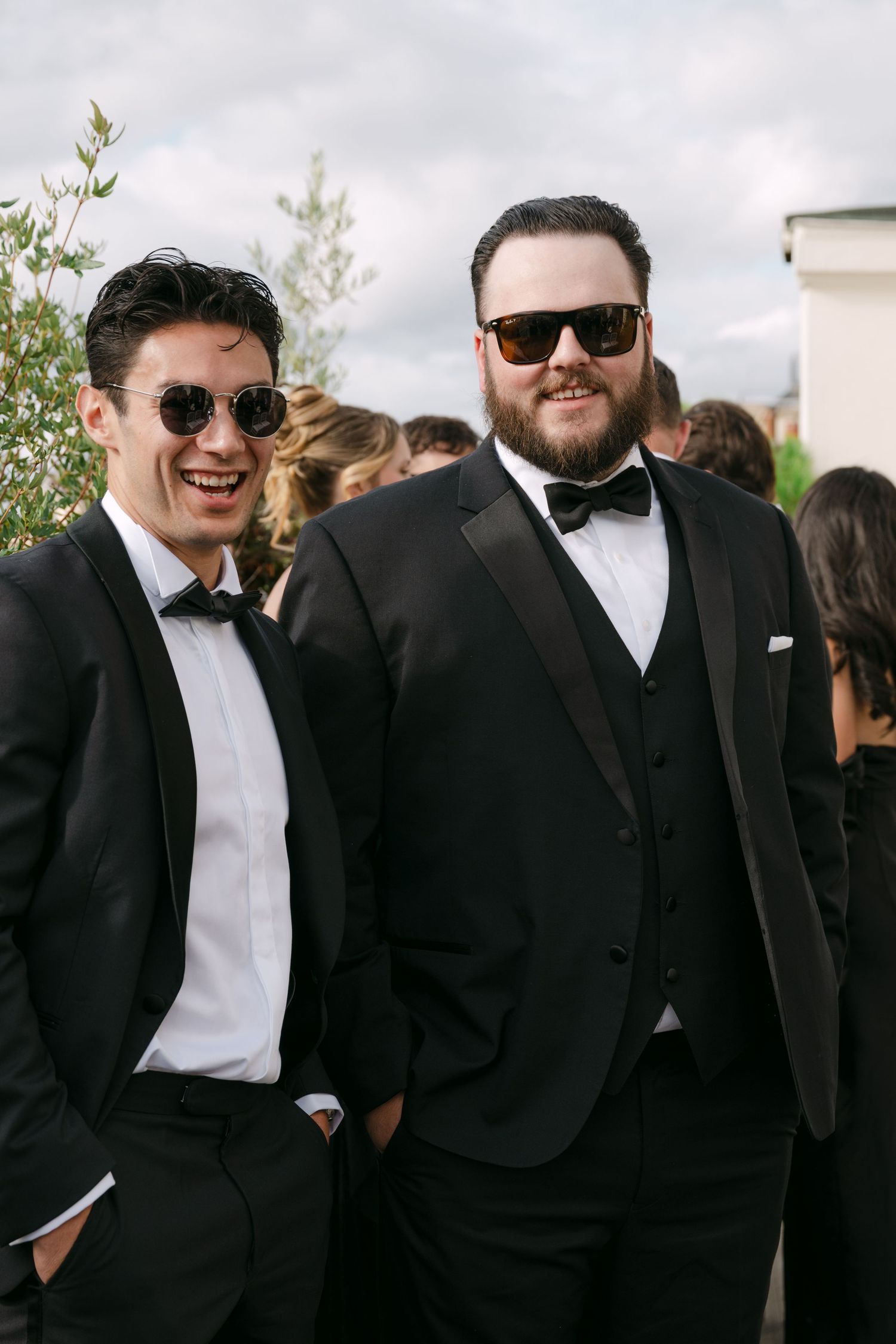 Groomsmen in black tuxedos and white bowties gather together at an outdoor wedding celebration.