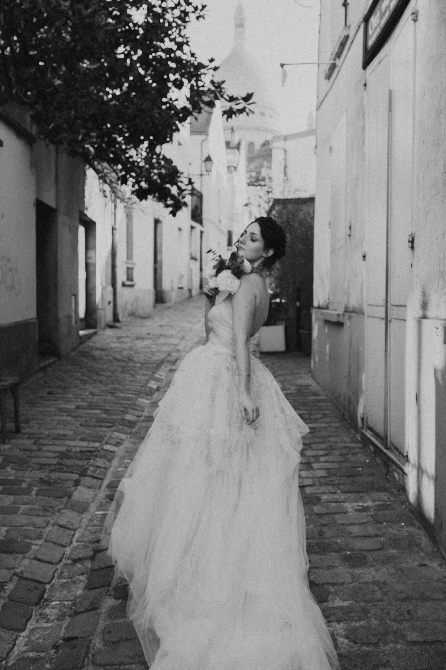 A black and white portrait of a bride in a ballgown twirling on a cobblestone street.