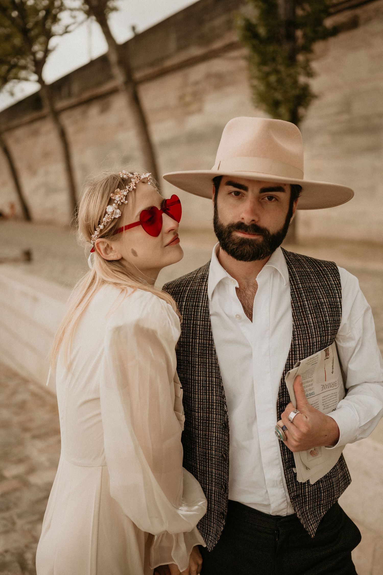 A stylish couple poses against a stone wall, wearing vintage-inspired outfits with a tan hat and red sunglasses.