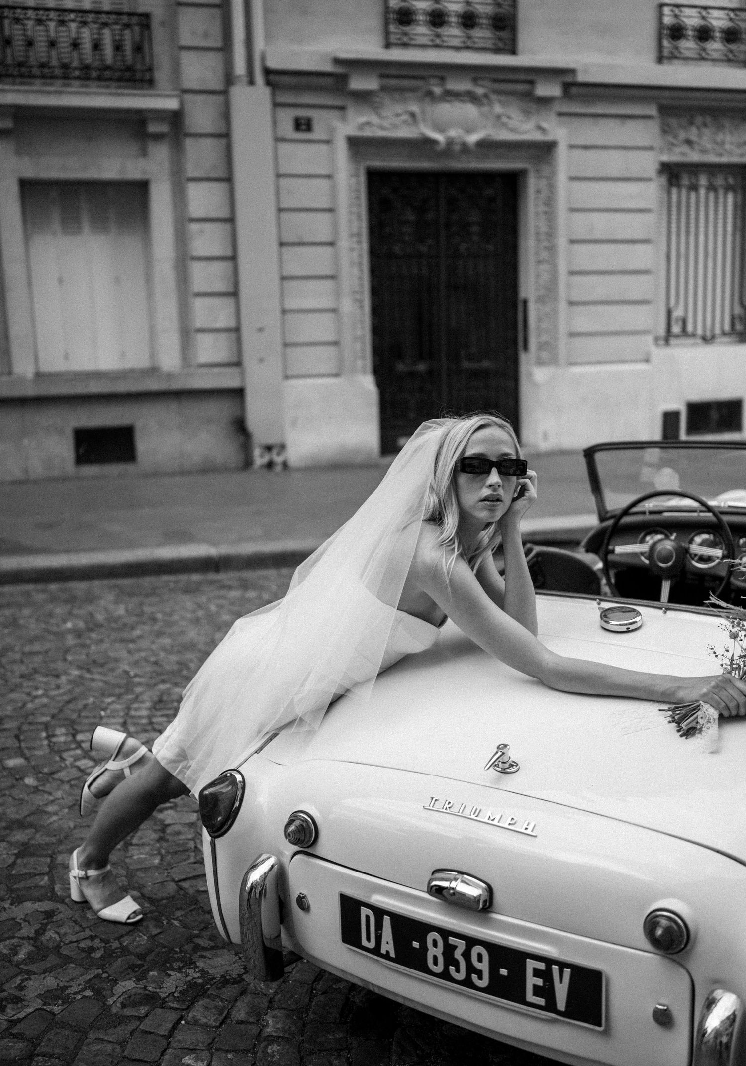 Black and white photo of bride in sunglasses leaning over vintage white Triumph TR3 convertible on Paris cobblestones.