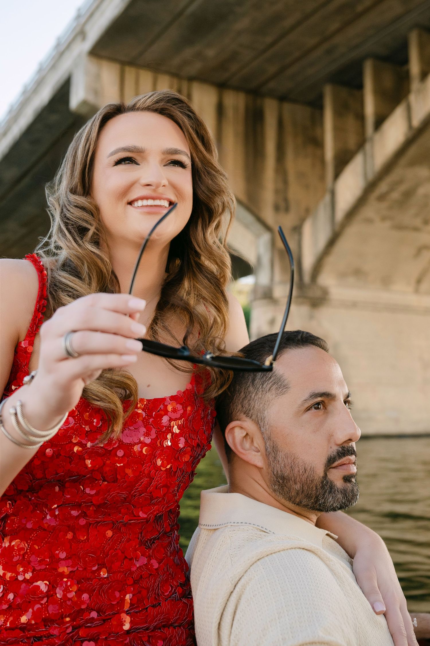 Smiling woman in red sequin dress holds sunglasses while embracing man under concrete bridge near river.