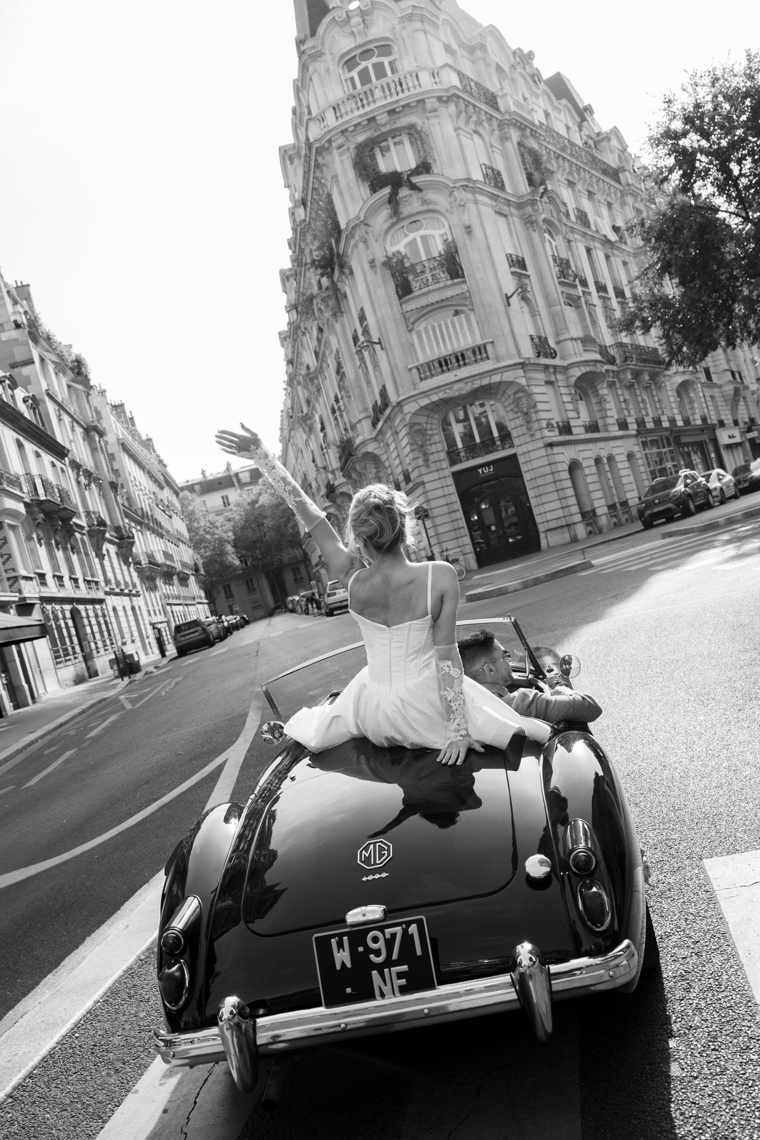 Black and white photo of bride sitting on vintage MG convertible waving through Parisian streets on wedding day.