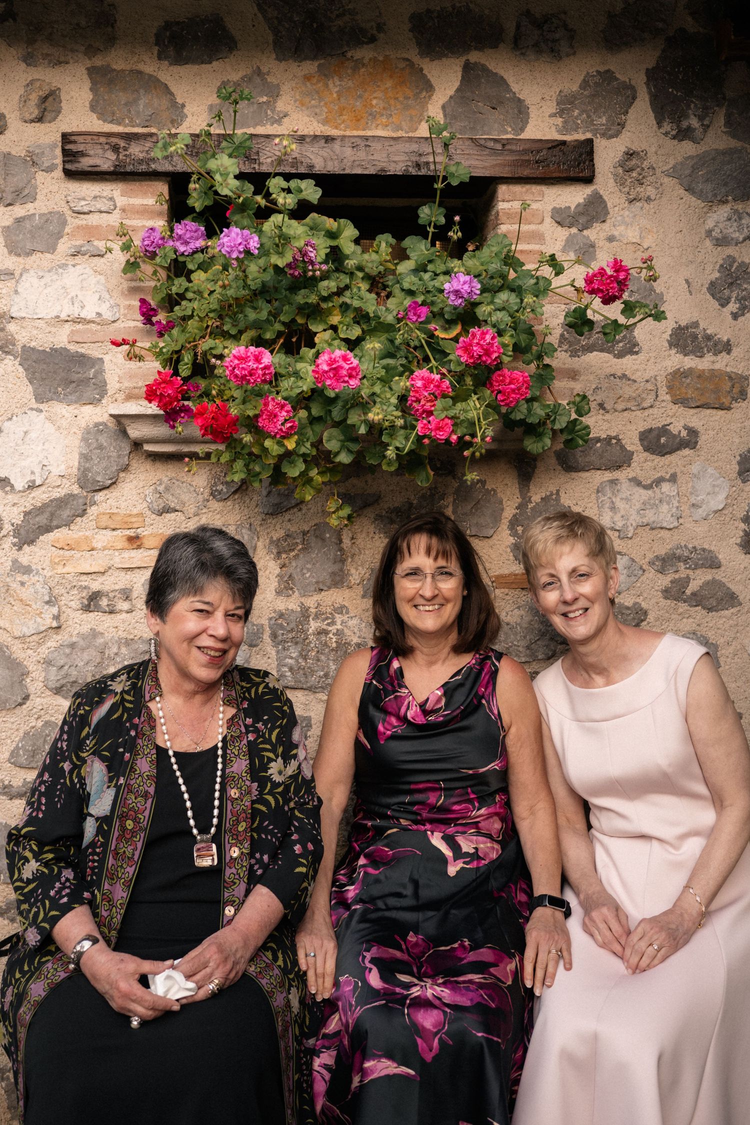 Three women smile together seated against a rustic stone wall adorned with vibrant pink geraniums.
