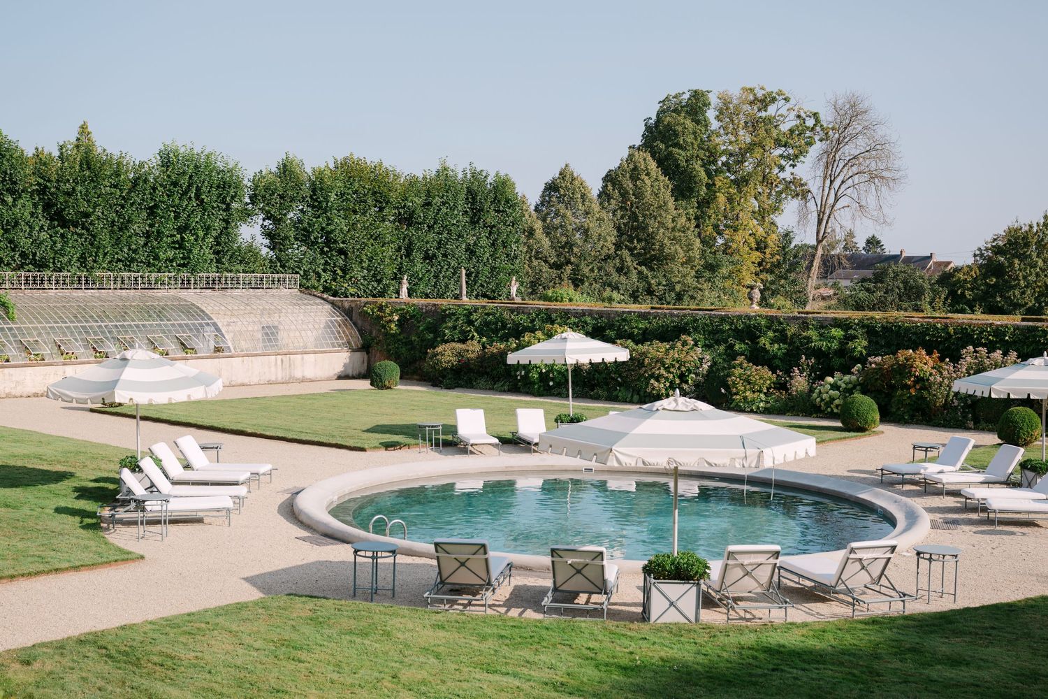 Aerial view of a circular luxury pool surrounded by white lounge chairs and umbrellas in formal garden grounds.