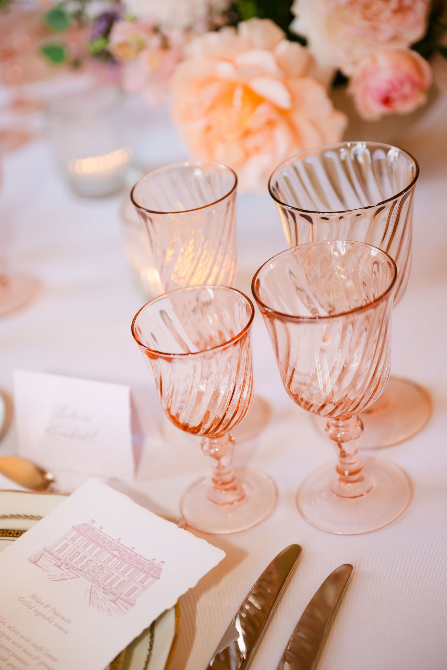 Four pink ribbed crystal wine glasses of varying heights arranged on a white wedding reception table.