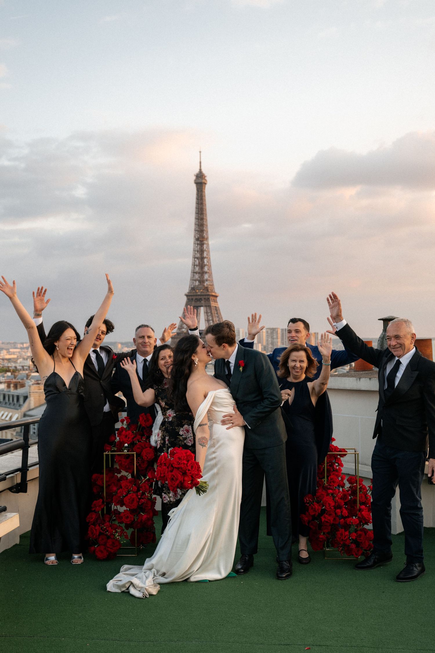 Newlyweds kissing on a Paris rooftop with Eiffel Tower backdrop surrounded by cheering wedding guests.