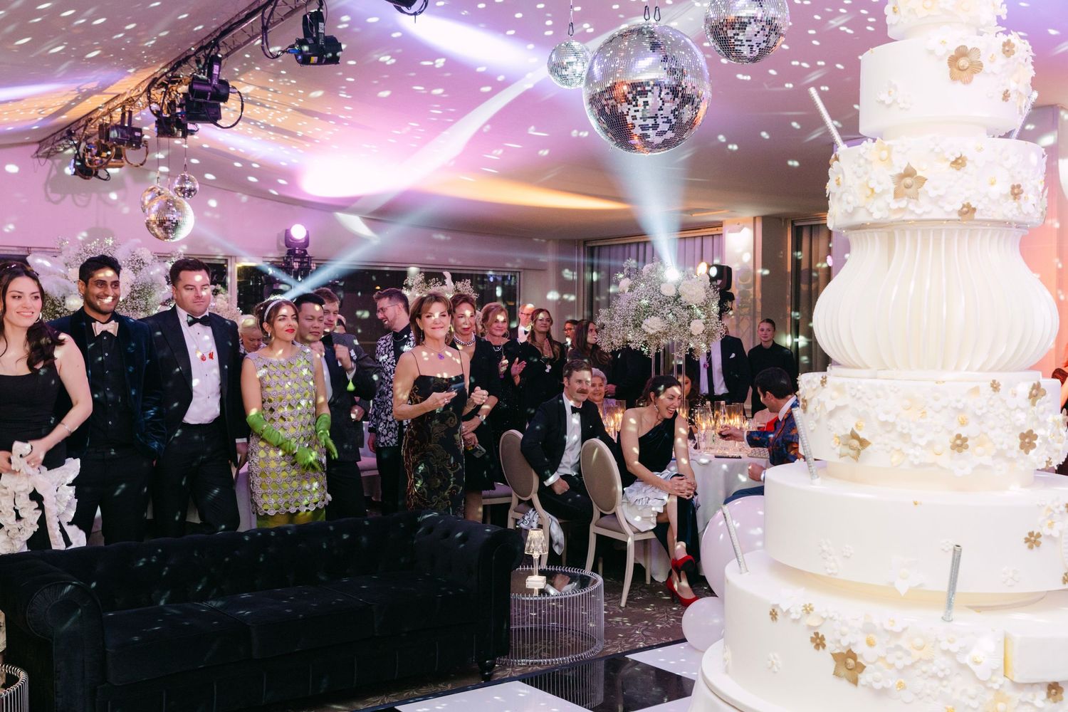 Wedding guests gathered around tall white gold-star cake on dance floor with disco balls above.