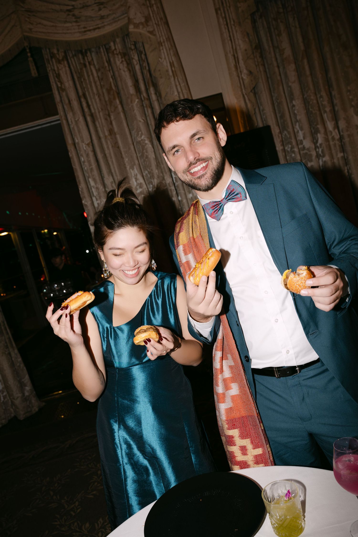 Smiling couple in formal attire holding mini tacos at an elegant wedding reception.