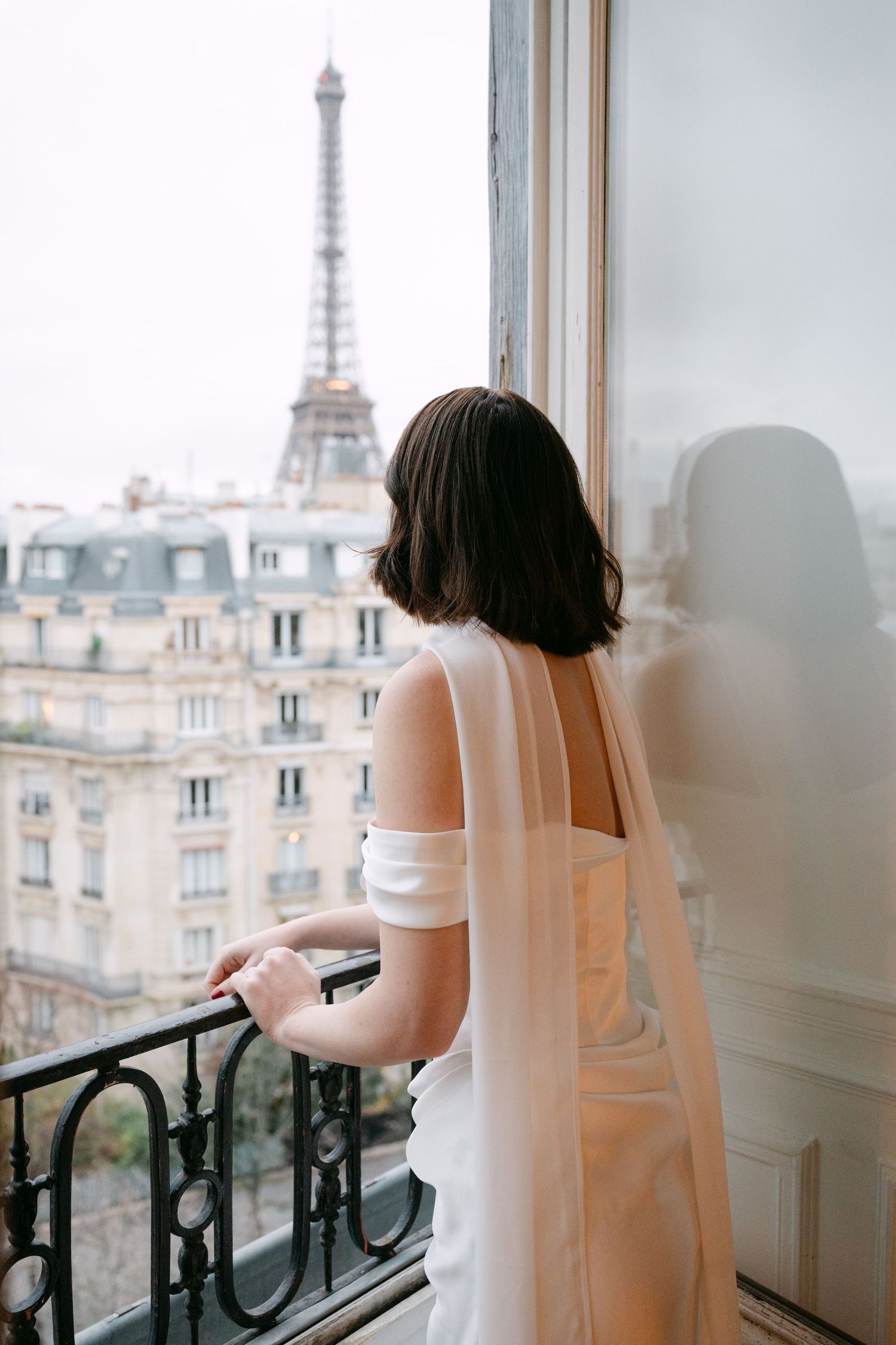 Bride in white gown with veil gazing at the Eiffel Tower from a Parisian hotel balcony.