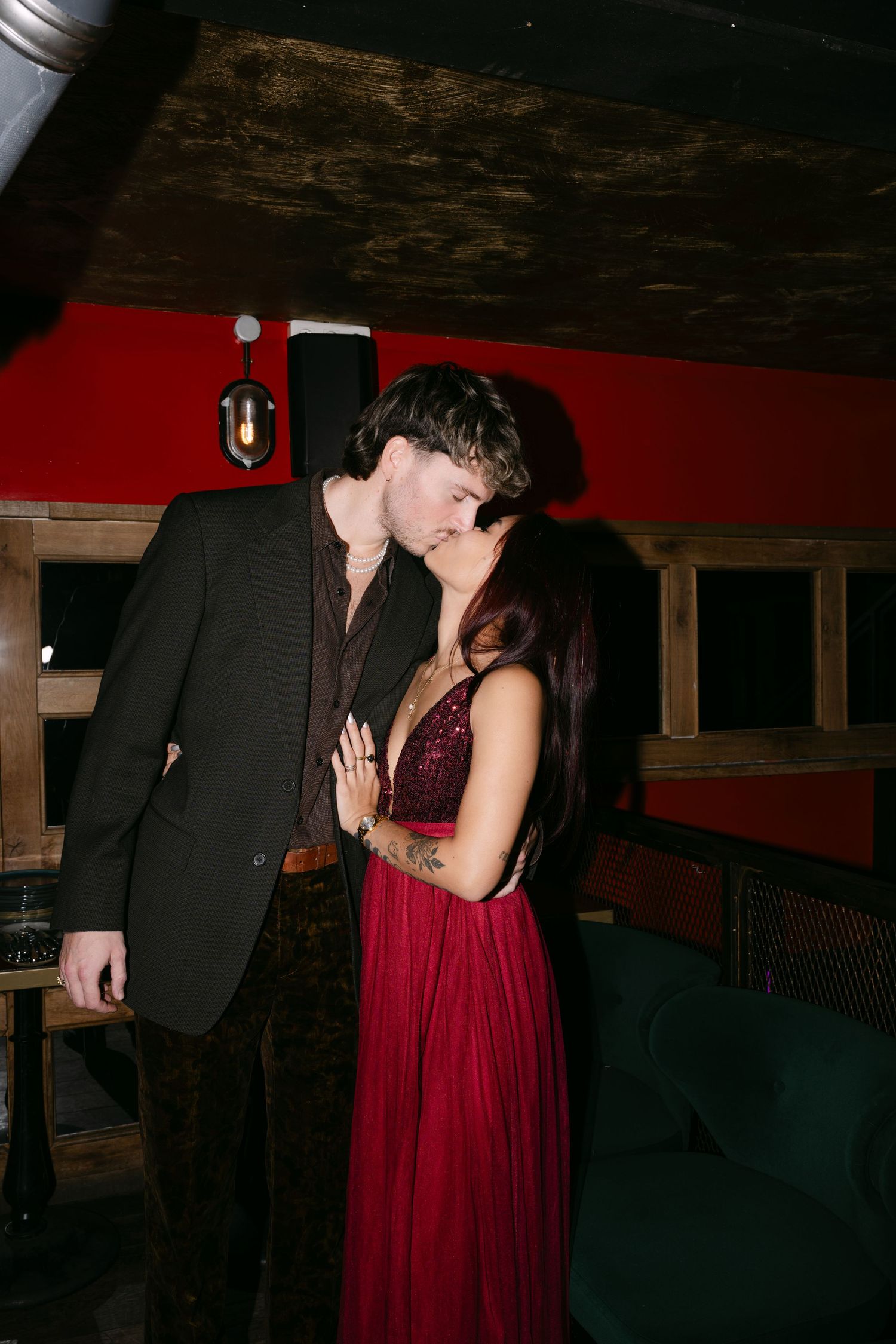 Couple sharing romantic kiss, woman in burgundy sequin dress in dimly lit venue.