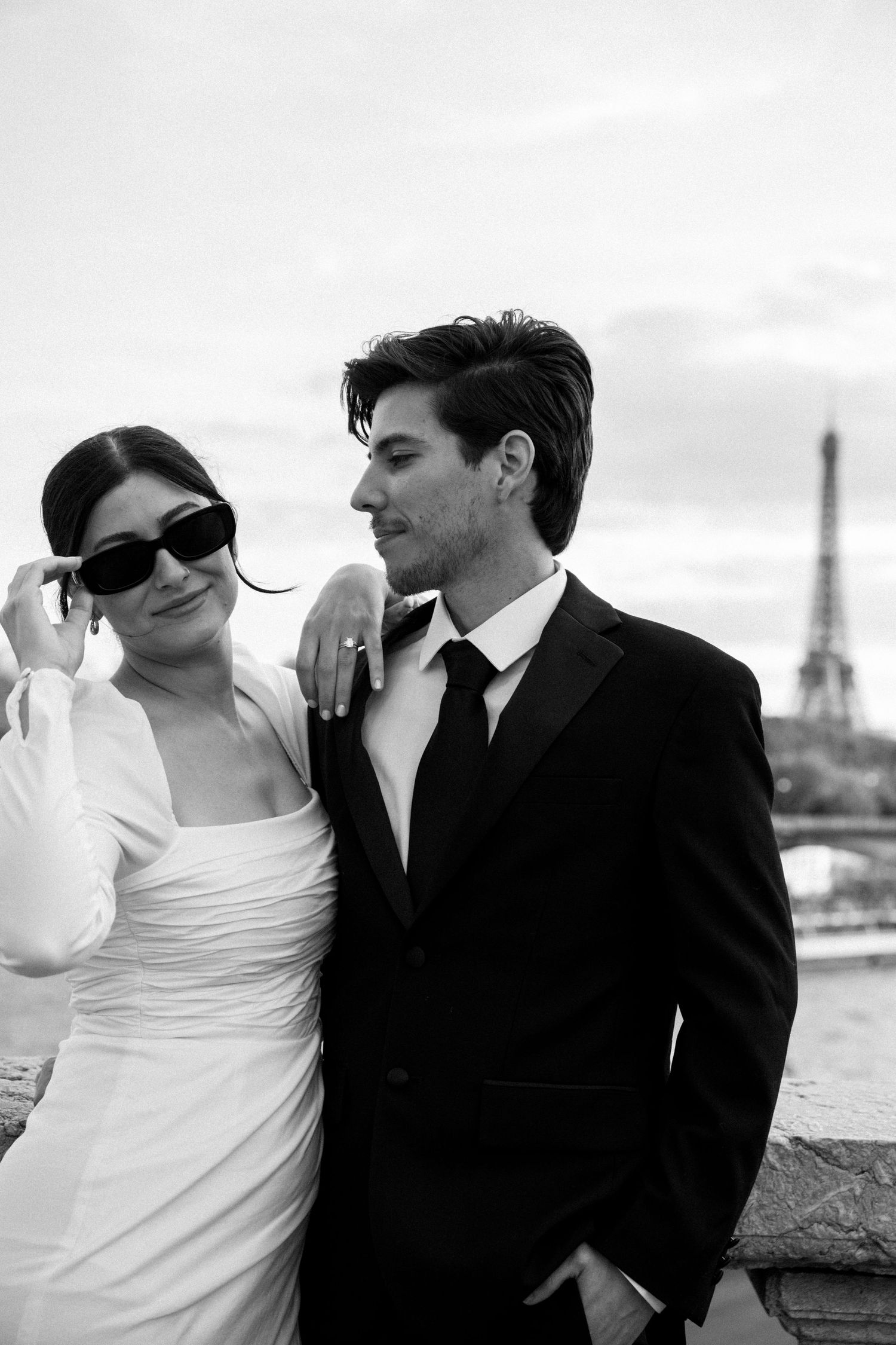 A stylish bride in sunglasses leans on her groom near the Eiffel Tower in a black and white photo.