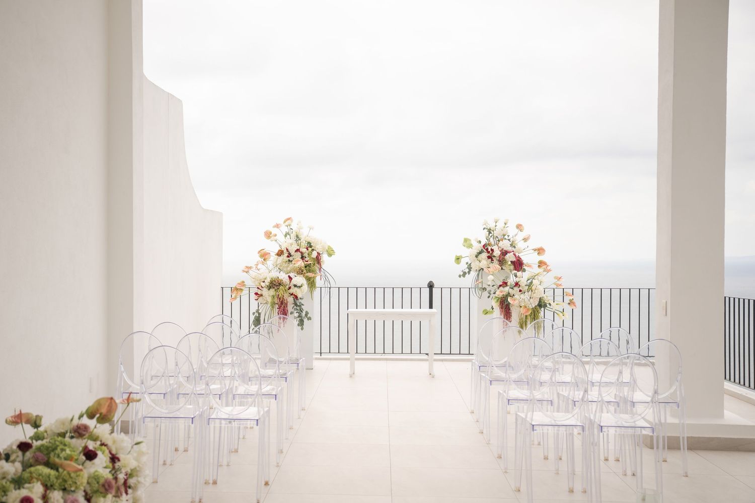 Elegant outdoor wedding ceremony setup with ghost chairs and floral arrangements on a white terrace overlooking the sea.