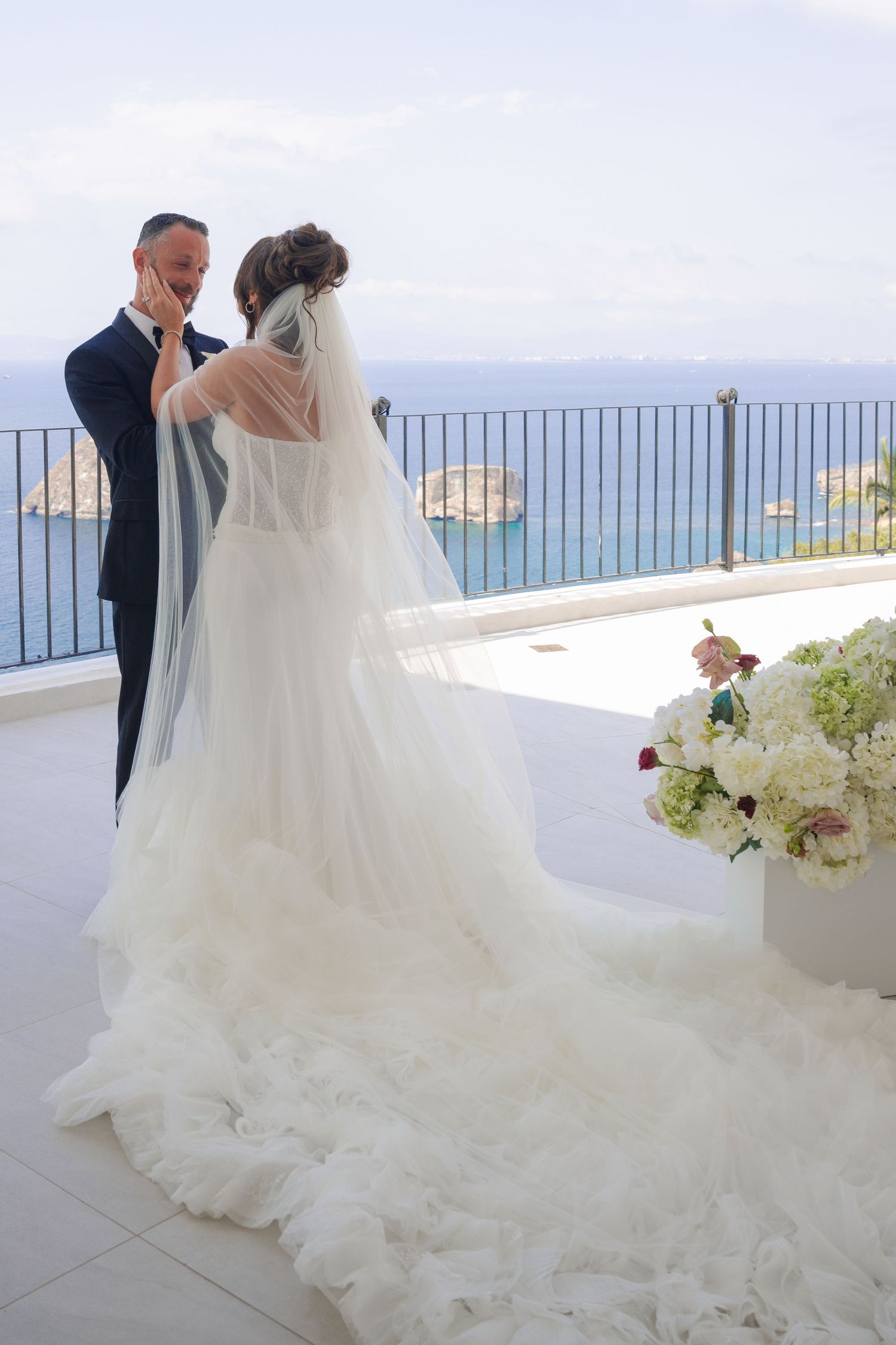 Bride and groom share a romantic moment on a terrace overlooking the sea, bride in a stunning feathered train wedding gown.