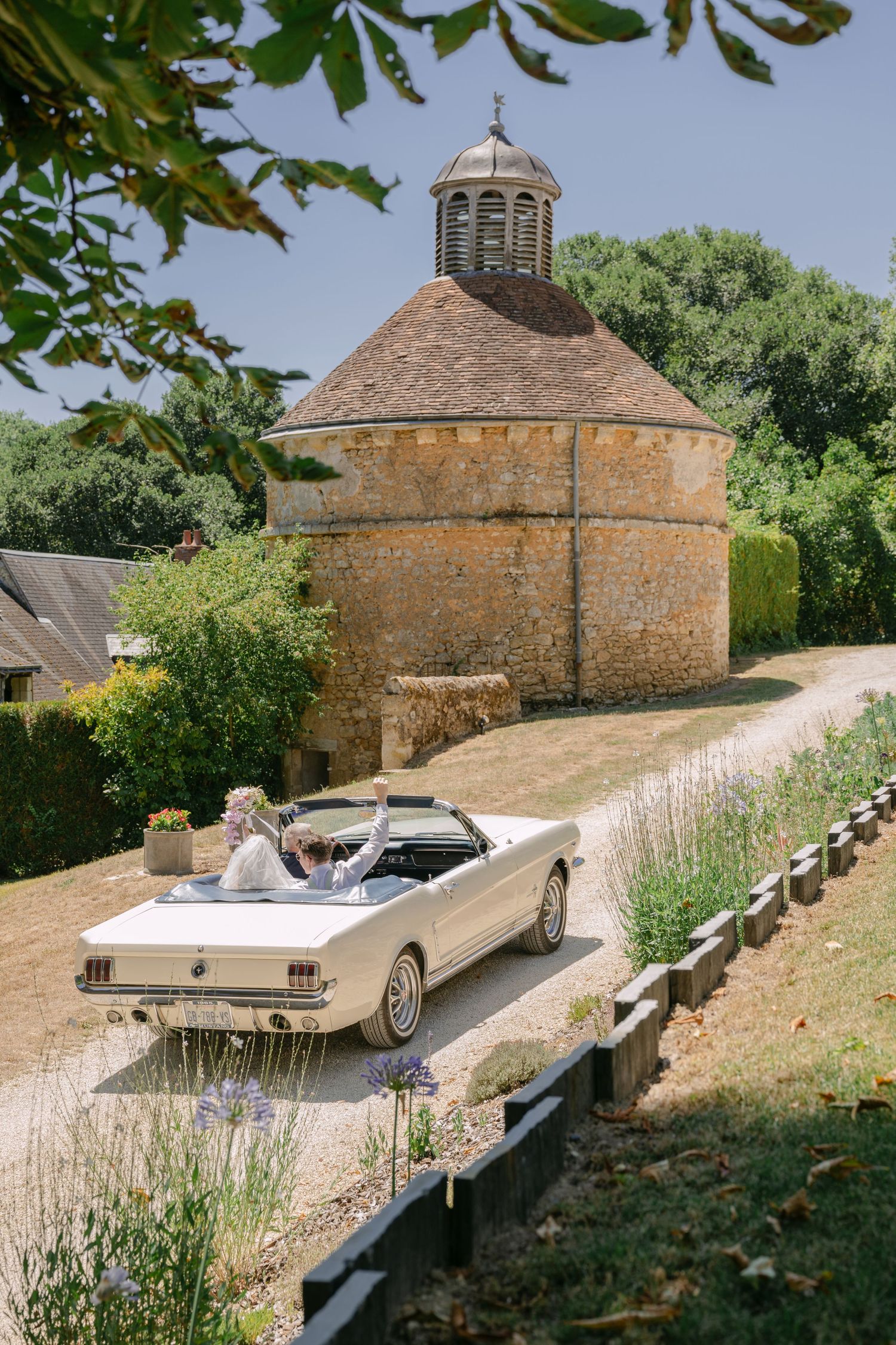 Bride and groom drive a classic white Ford Mustang convertible past a historic stone dovecote in the English countryside.