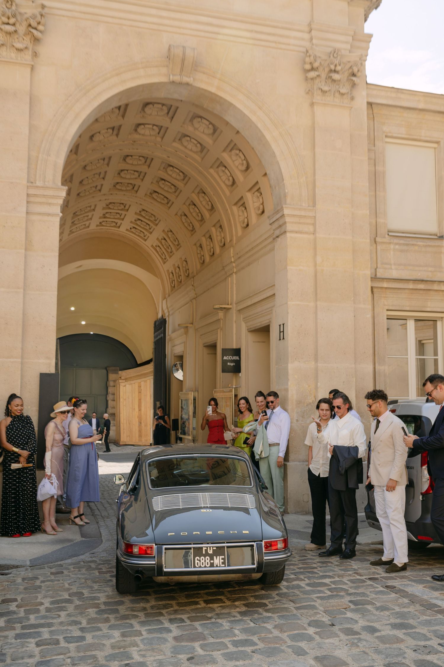 Vintage Porsche parked under grand stone arch in Paris, surrounded by elegantly dressed guests at outdoor event.