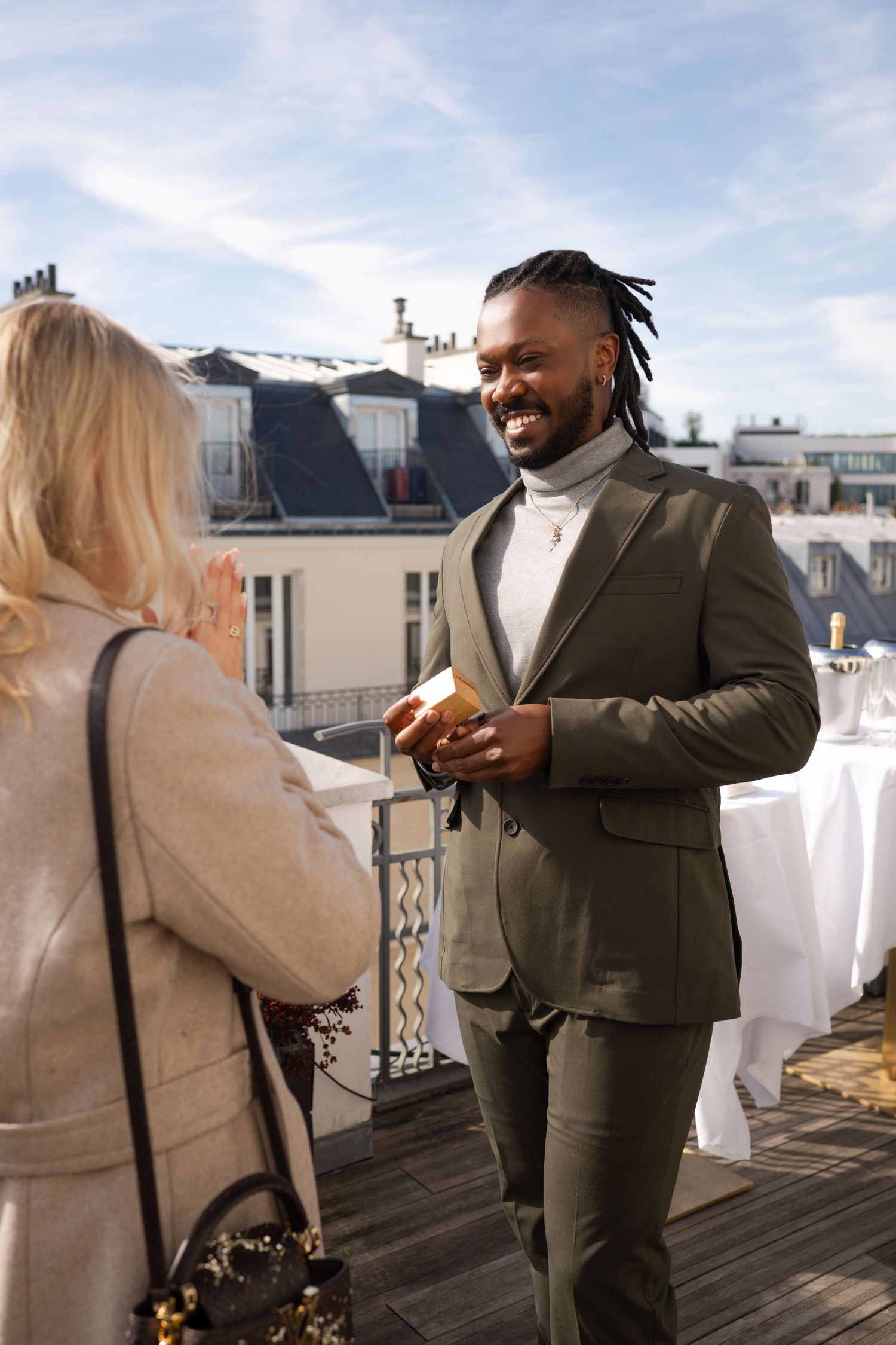 A smiling man in an olive suit holds a gift box while chatting with a blonde woman on a sunny Parisian rooftop terrace.