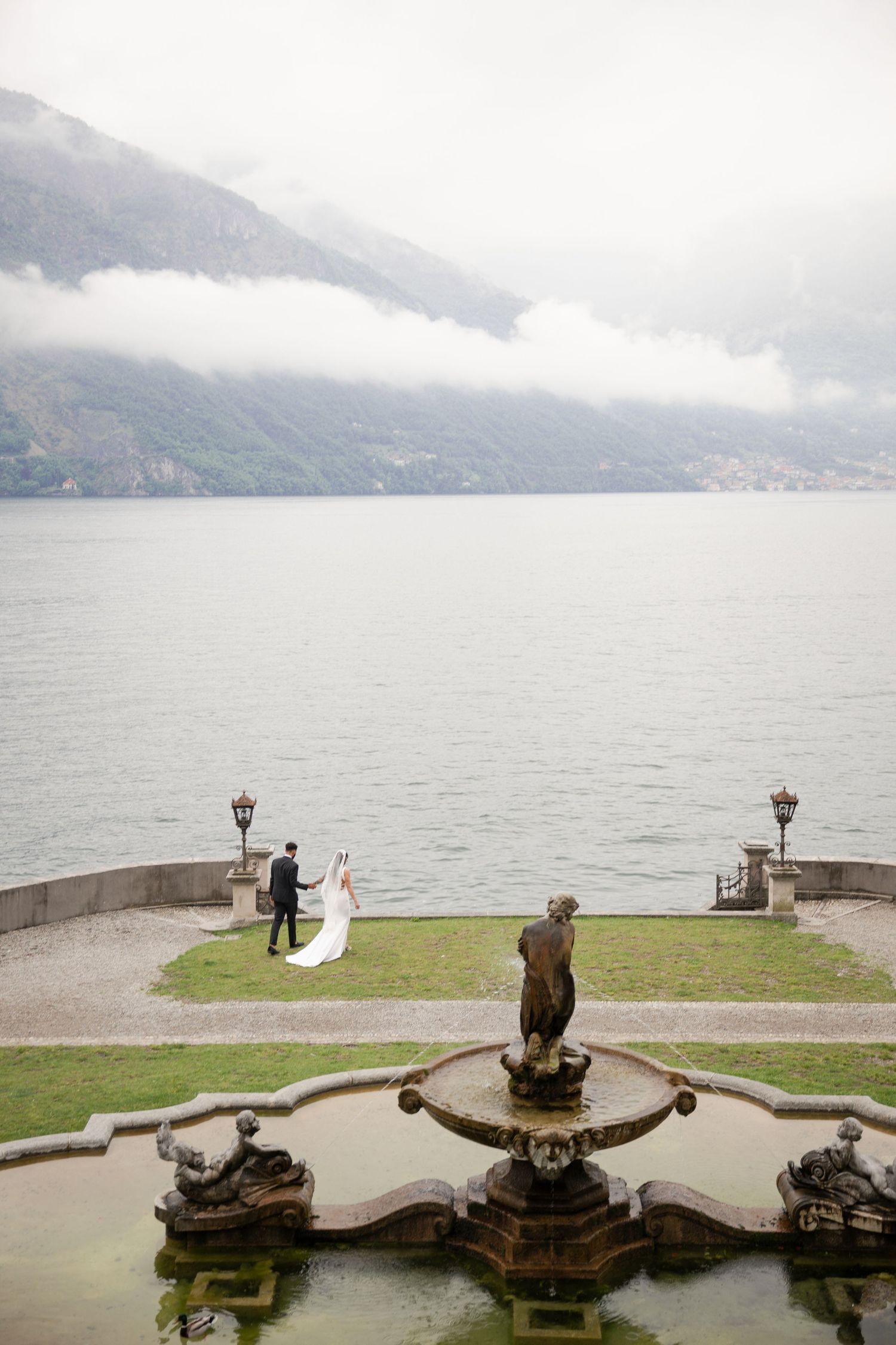 Aerial view of bride and groom holding hands near ornate fountain with misty Lake Como mountains behind them.