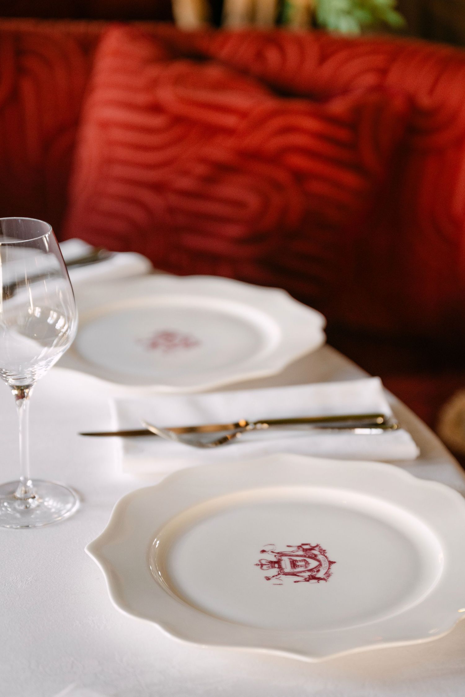 Elegant restaurant place setting featuring monogrammed white porcelain plates, crystal wine glass, and gold cutlery.