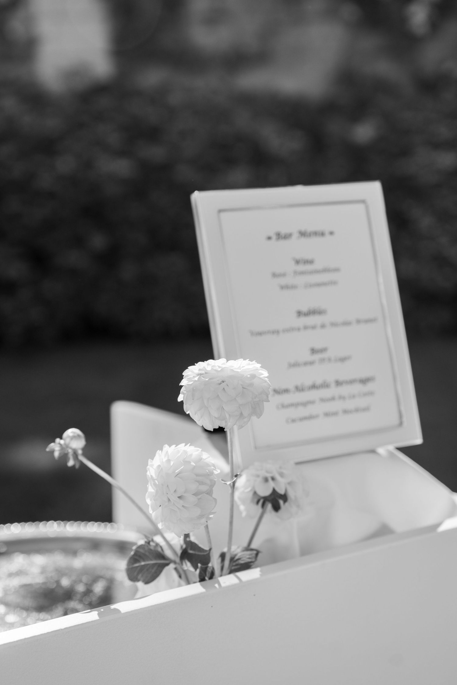 Black and white photo of white flowers beside a bar menu card at an outdoor wedding reception.