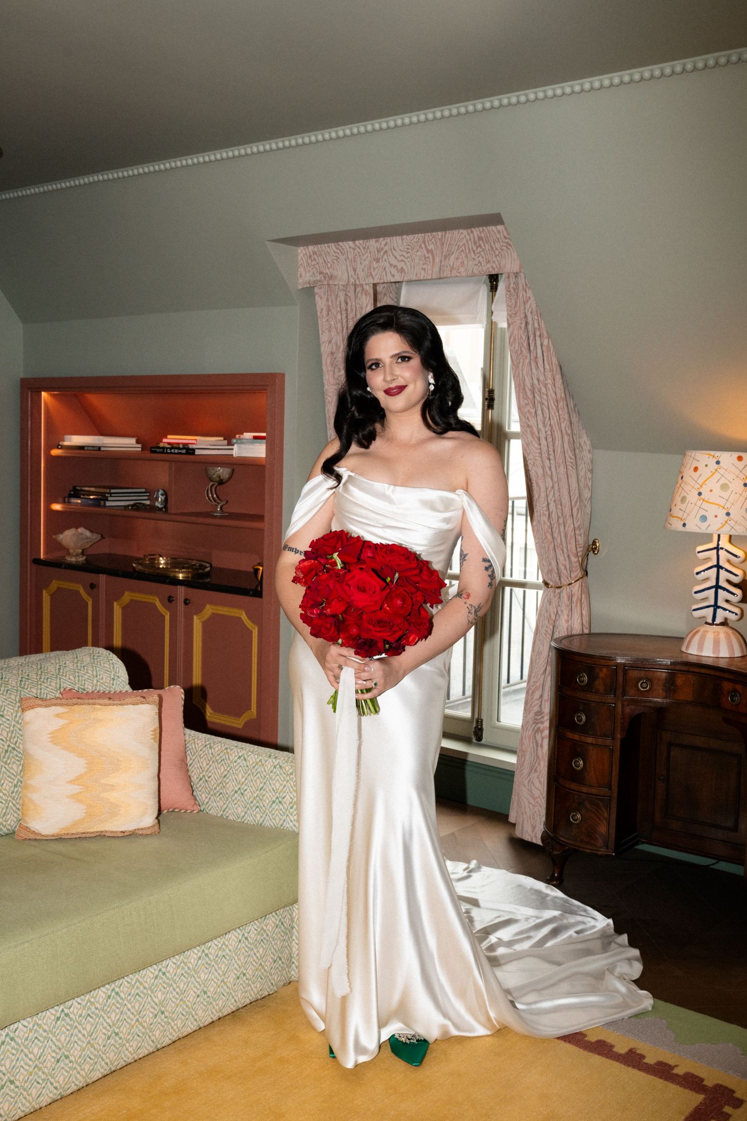 Stunning bride in off-shoulder white satin gown holds vibrant red rose bouquet while standing in elegant hotel suite.
