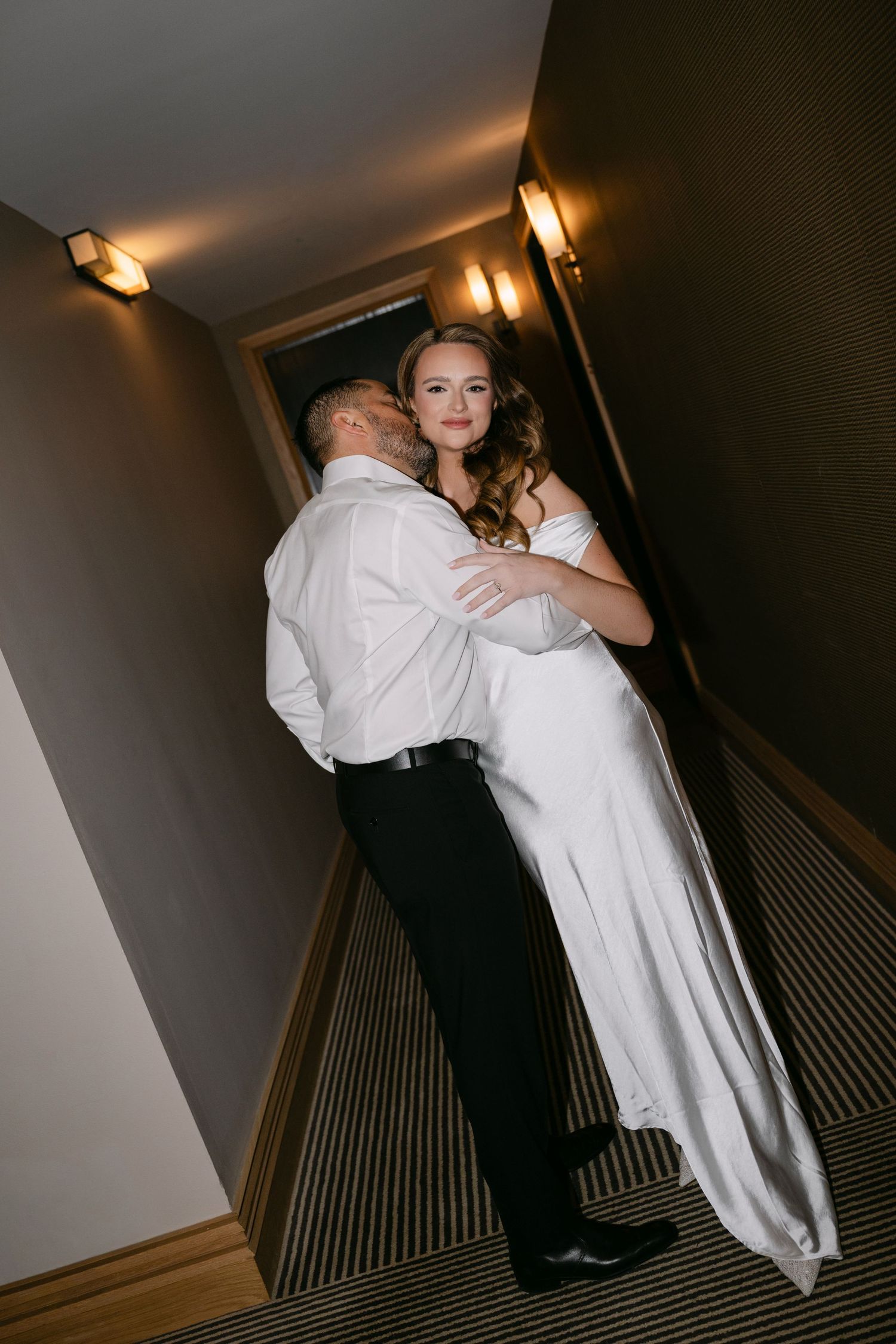 Groom embracing bride from behind in dimly lit hotel corridor, bride smiling at camera in white gown.