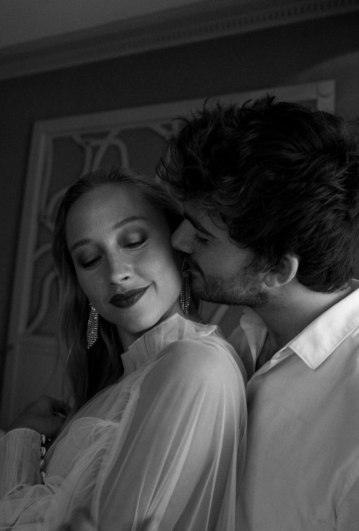 Black and white photo of a couple sharing an intimate moment, man kissing womans cheek as she smiles softly.