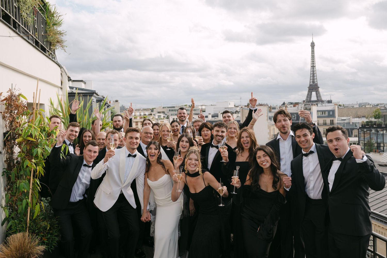 Large wedding party group photo on a Paris rooftop terrace with the Eiffel Tower visible in the background.
