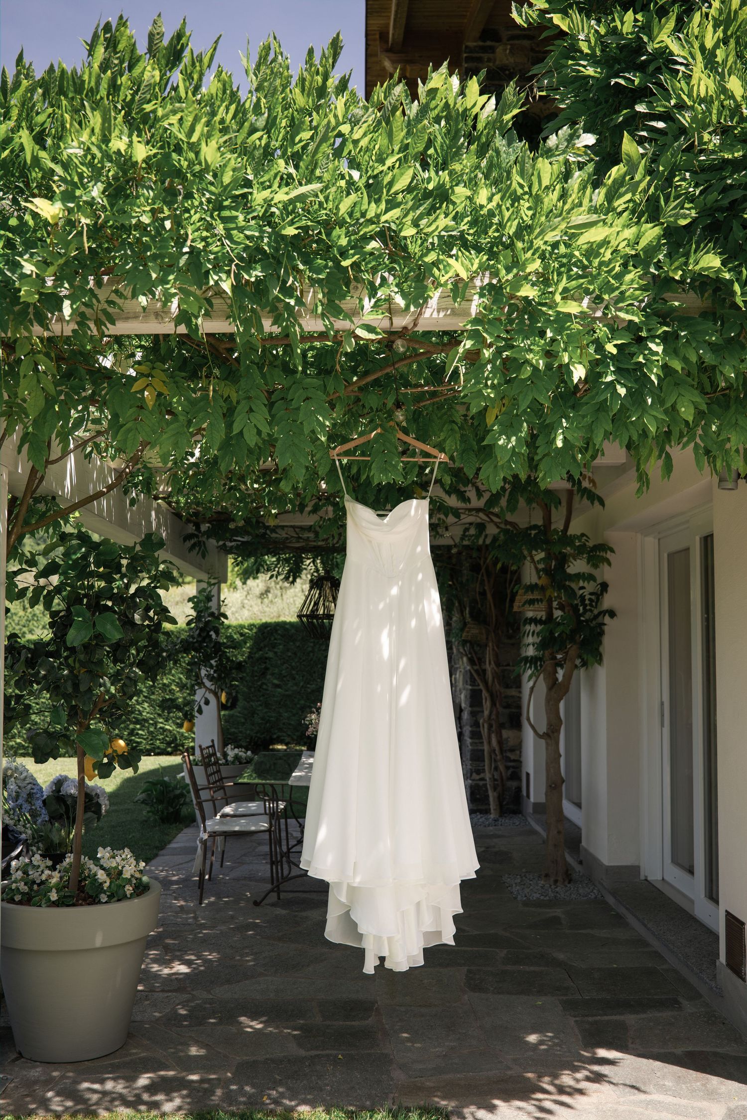White wedding dress hanging on a wooden hanger under a lush green pergola in a sunlit outdoor garden setting.
