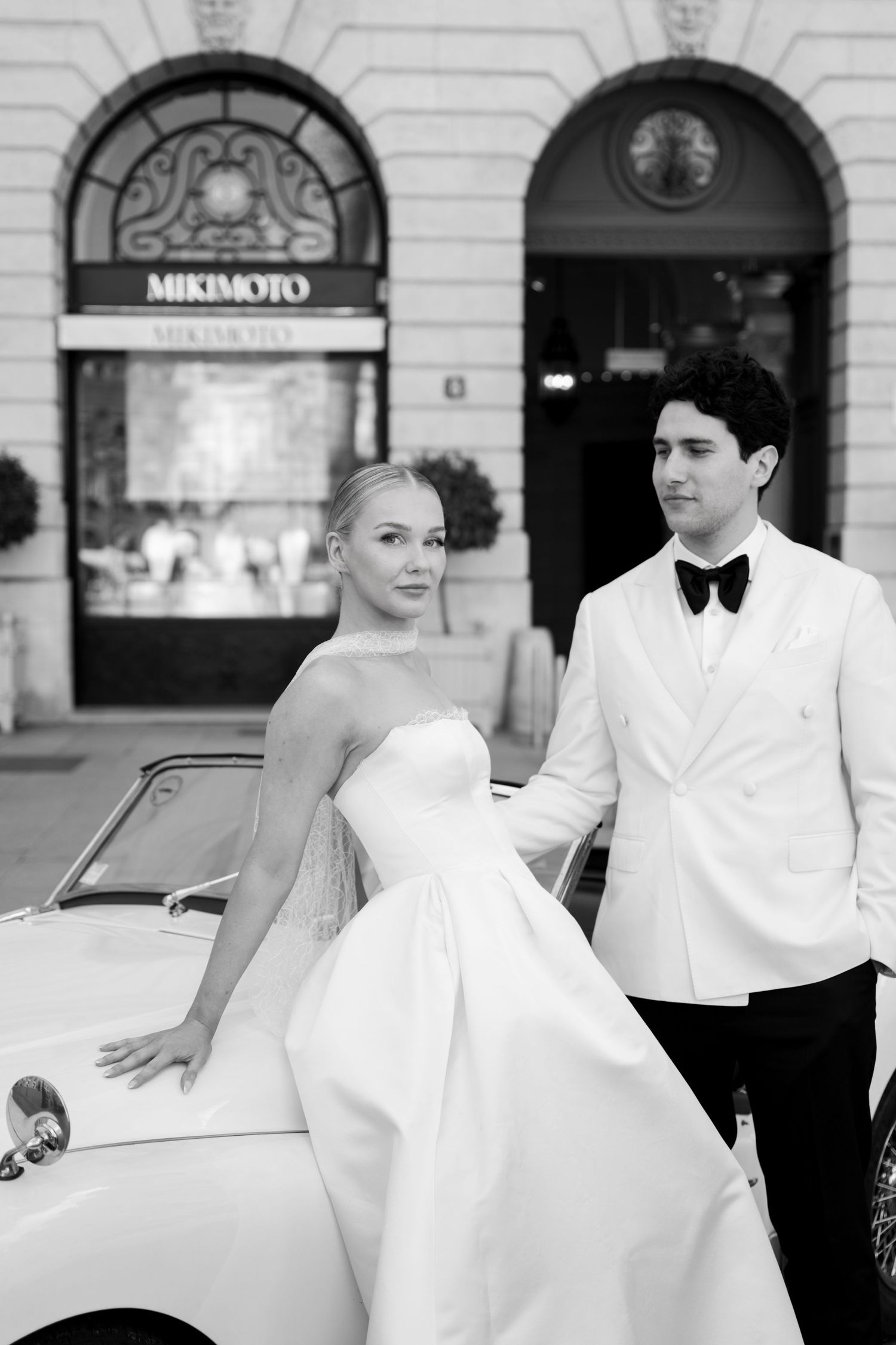 Elegant bride and groom in black and white photo posing beside vintage car outside Mikimoto store.