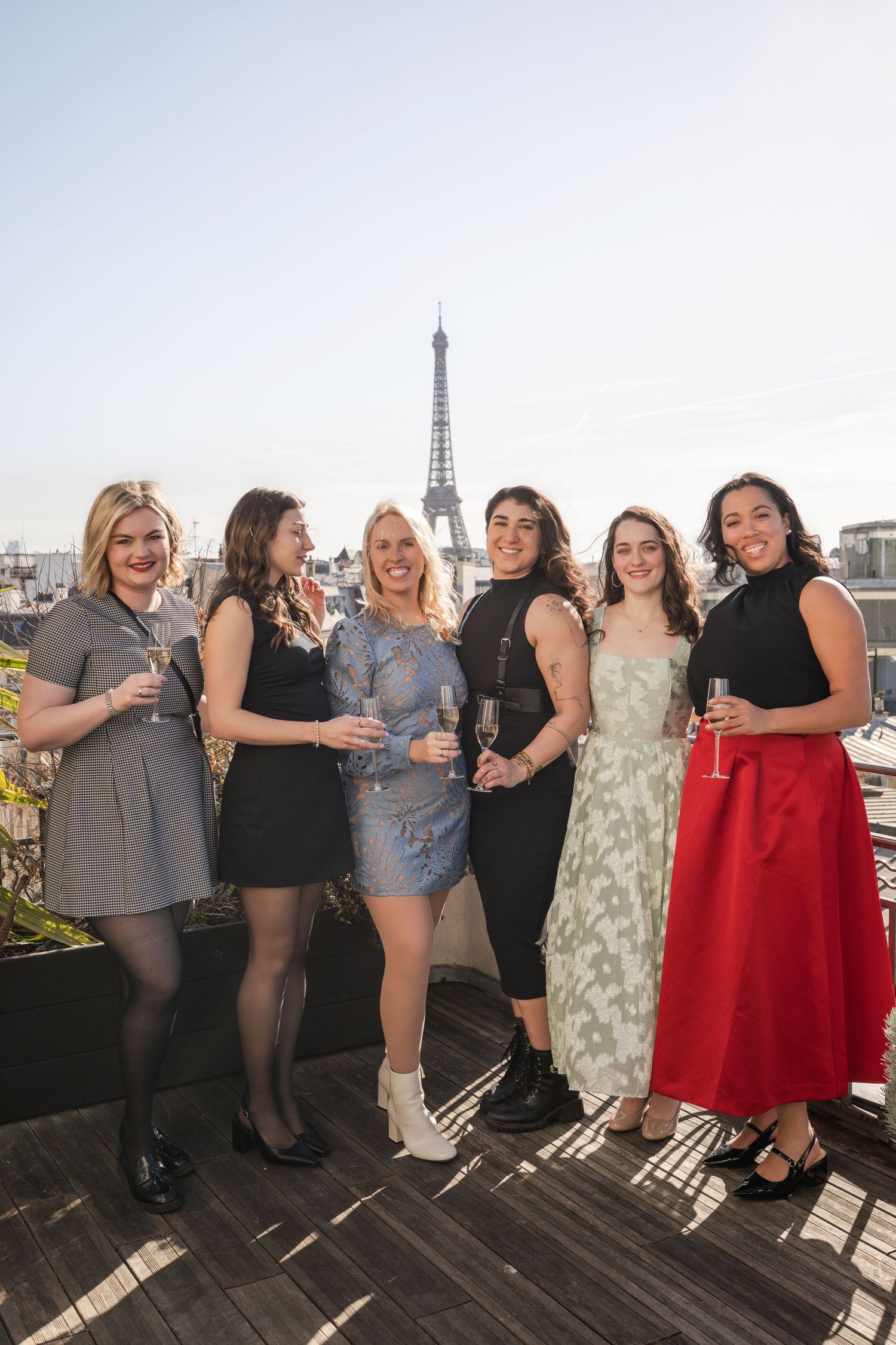 Group of six women posing with champagne on Paris rooftop with Eiffel Tower backdrop.
