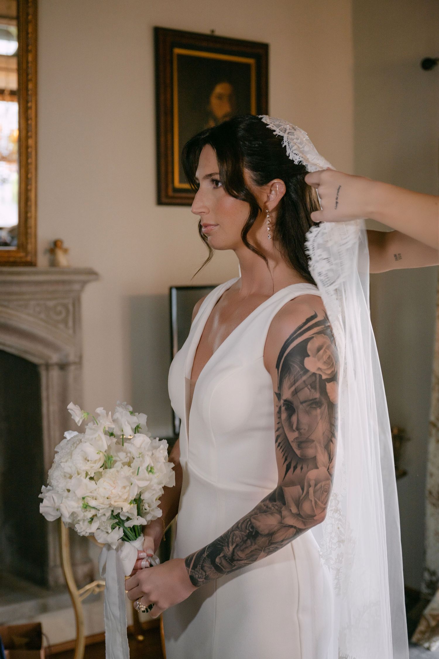 Tattooed bride in white gown holds bouquet of white flowers, wearing veil and lace headpiece indoors.