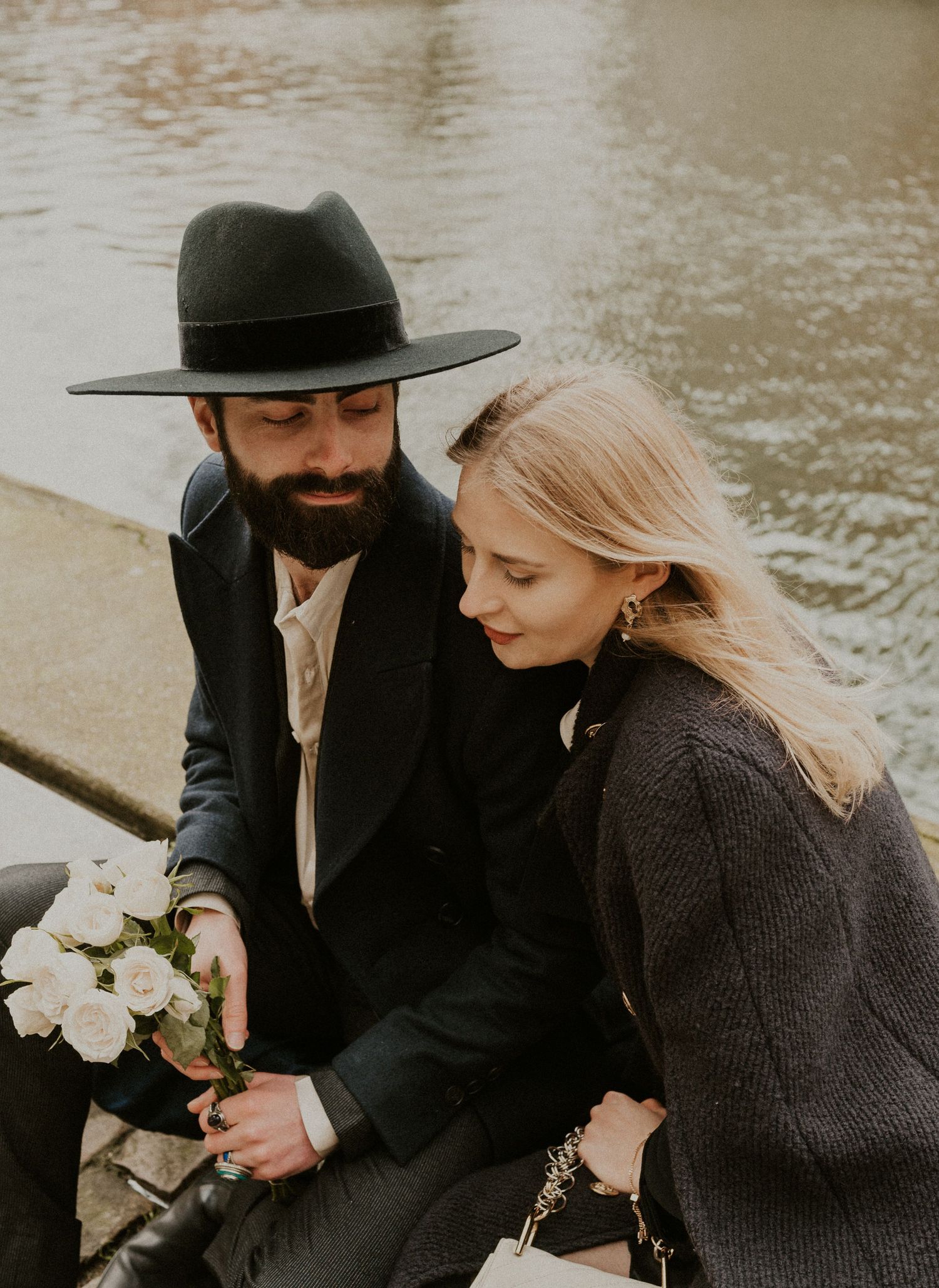 Stylish couple in dark winter coats share intimate moment with white flowers by water.