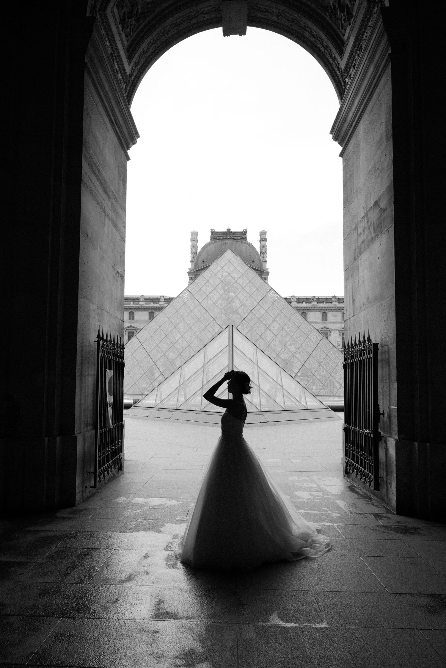 Silhouette of bride in ballgown against Louvre pyramid archway in Paris.