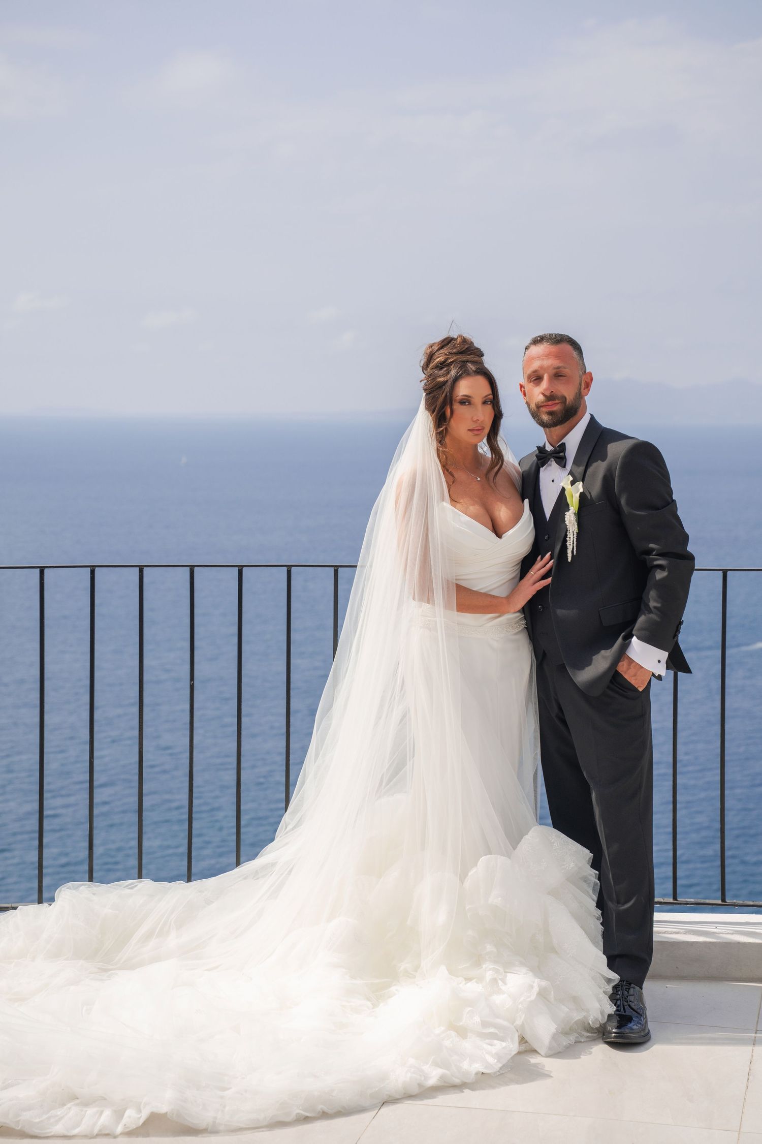 Bride and groom pose together on oceanfront balcony, she in ruffled gown, he in black tux.