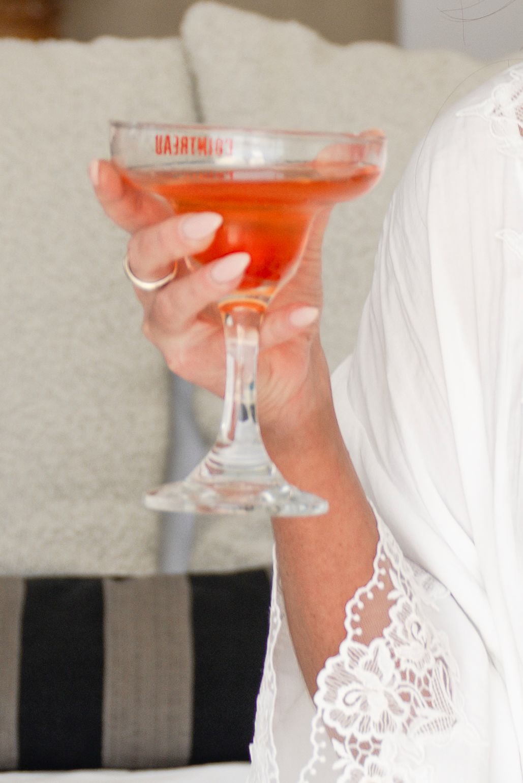 Woman in white lace robe holding a pink cocktail in a margarita glass while sitting on a couch.