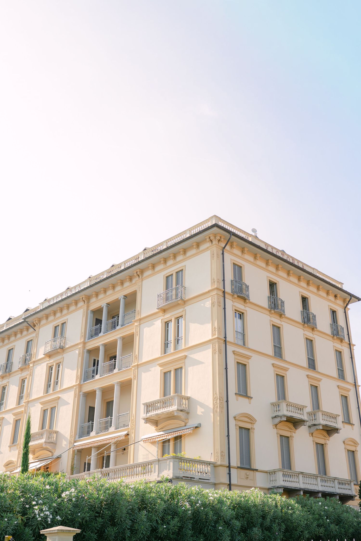 Elegant cream-colored European hotel building with ornate balconies and lush green hedges under a soft sky.