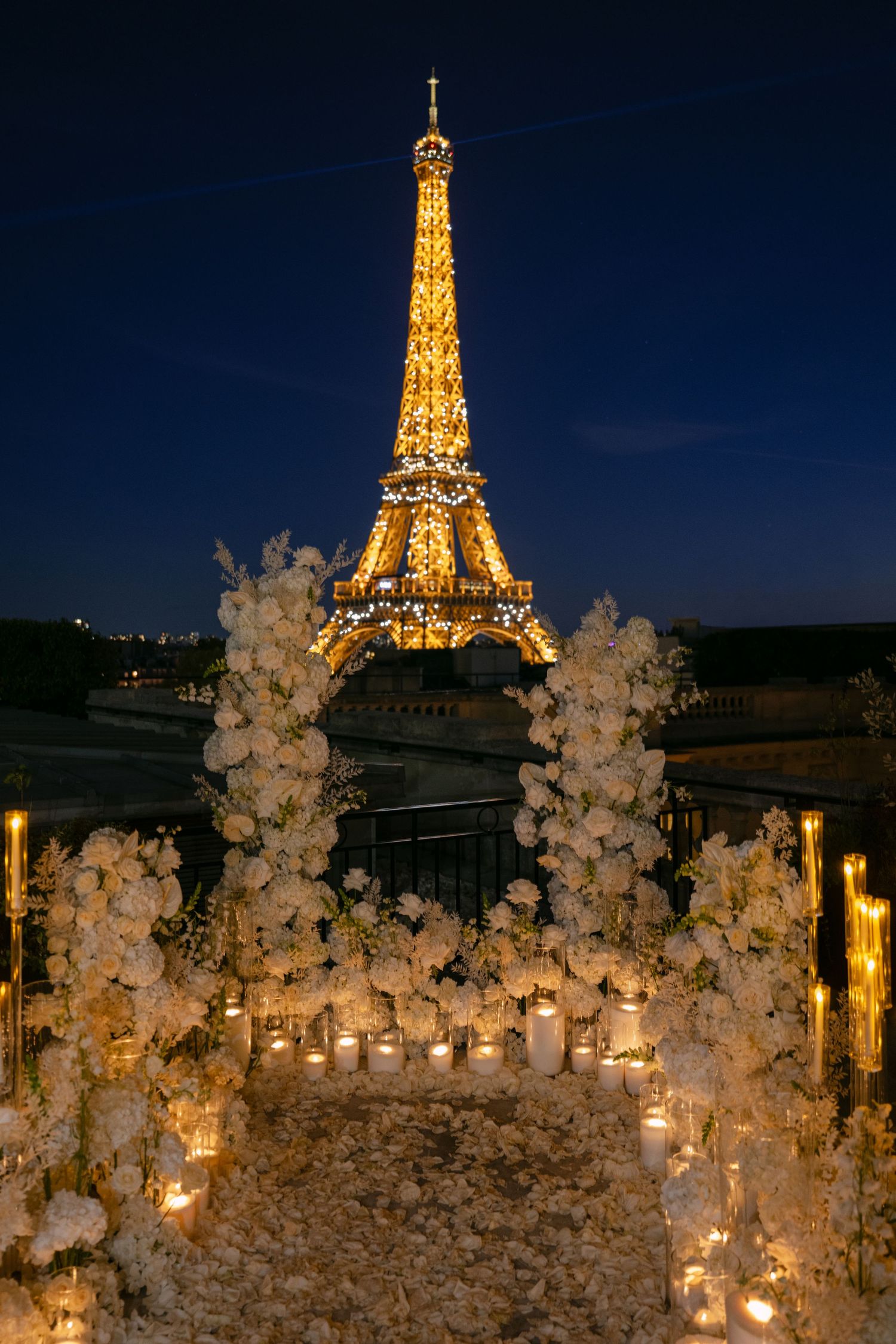 Illuminated Eiffel Tower glows at night behind white floral arrangements and glowing candles on a Paris proposal terrace.