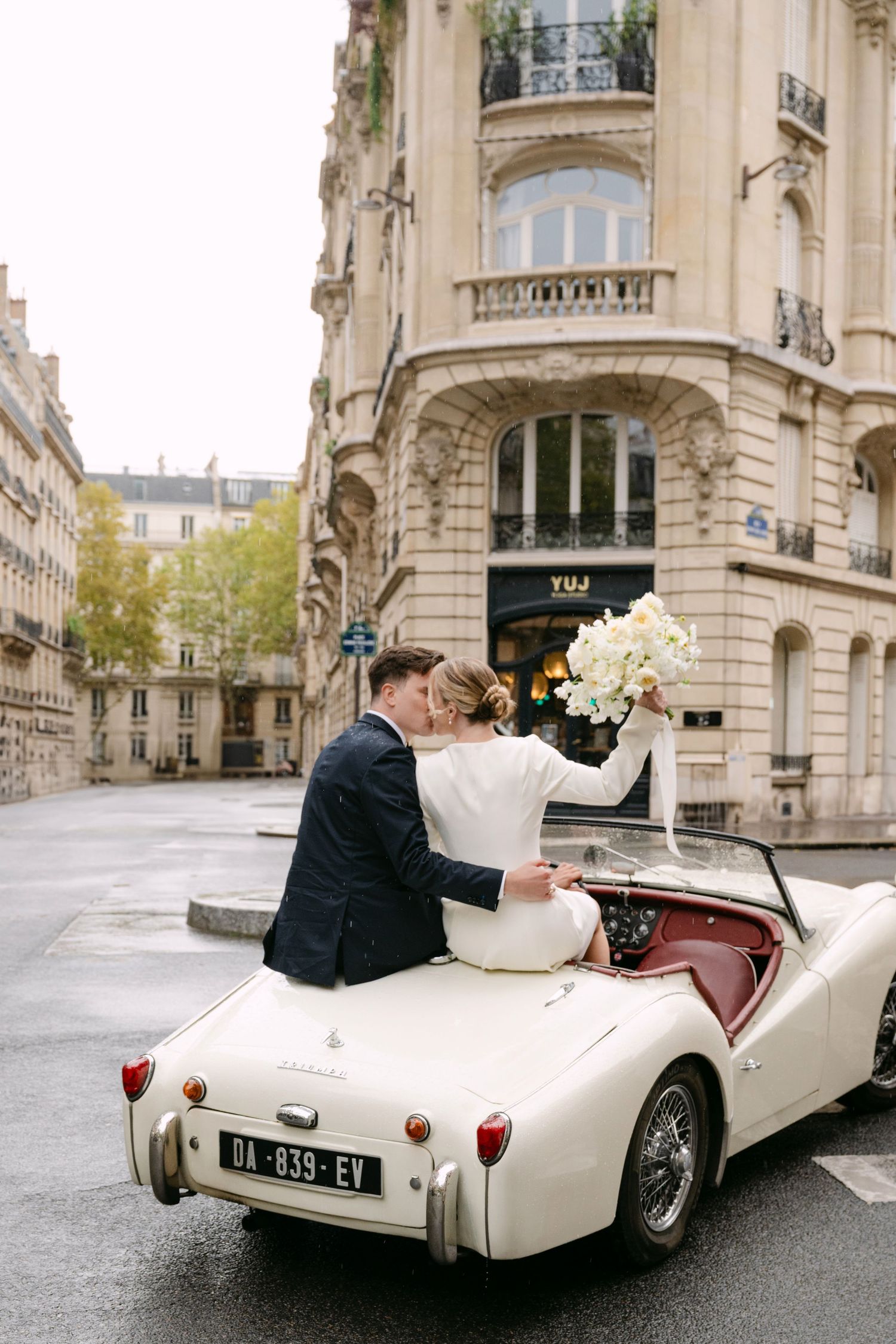Newlyweds kiss in a vintage white convertible on a Parisian street, bride holding white floral bouquet aloft.
