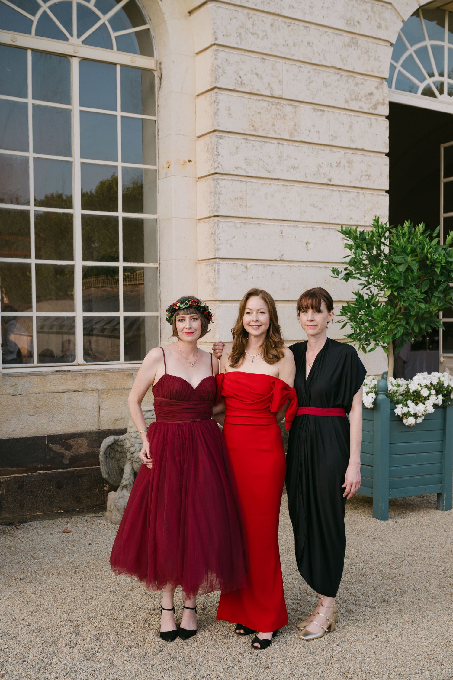 Three women in red and black formal dresses posing outside a stone building.