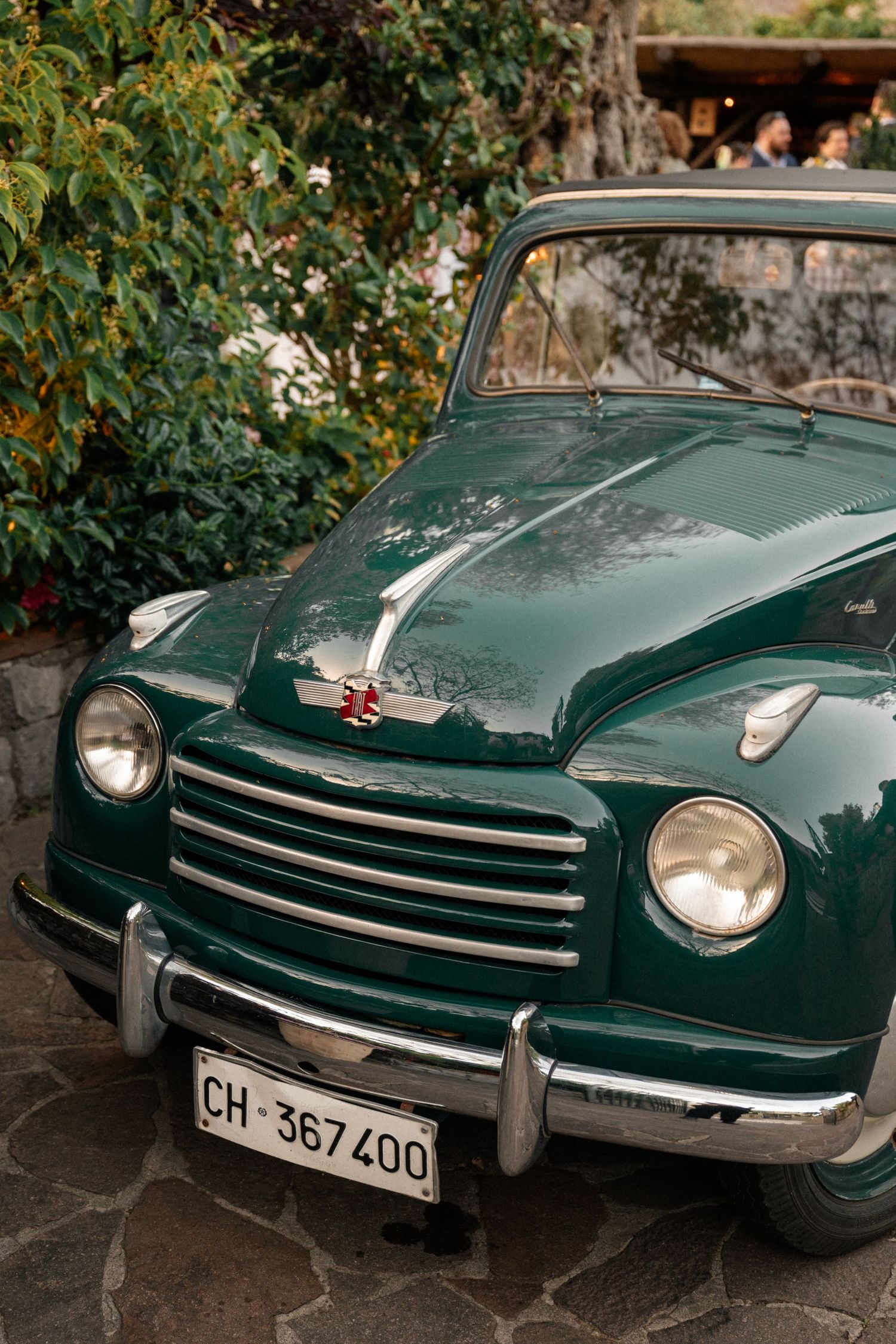 Vintage green Fiat 500 classic car with chrome details parked on stone driveway surrounded by greenery.