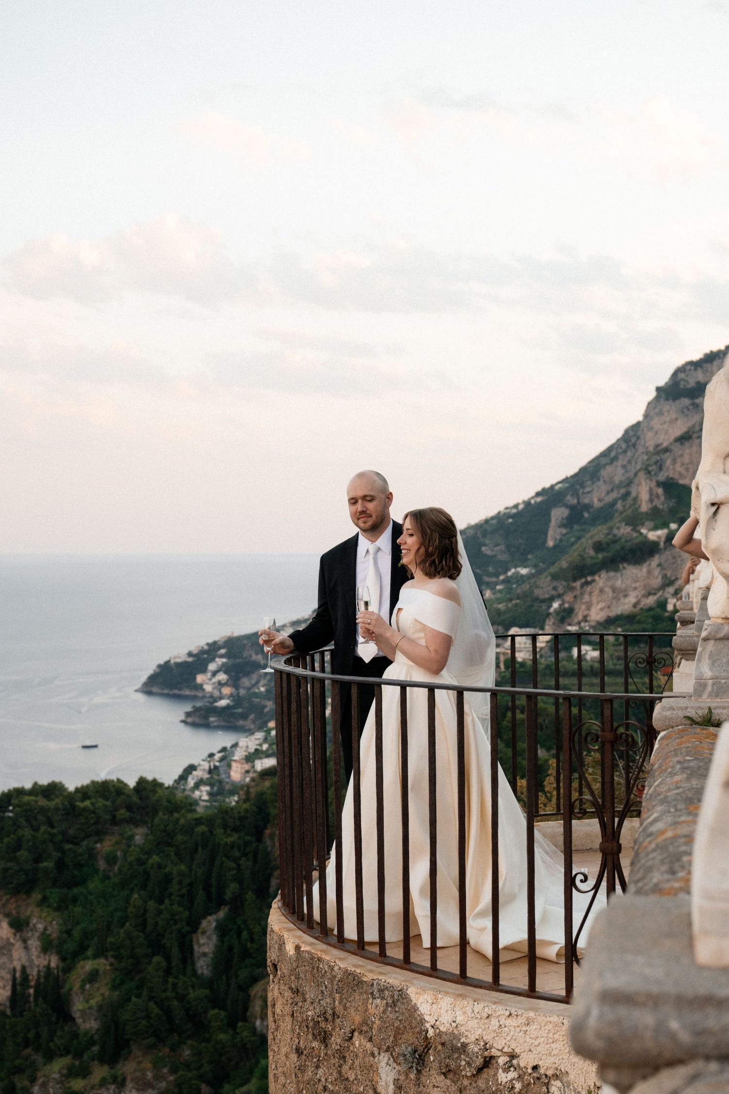 Newlyweds share champagne on scenic balcony overlooking Italian coastline at sunset.