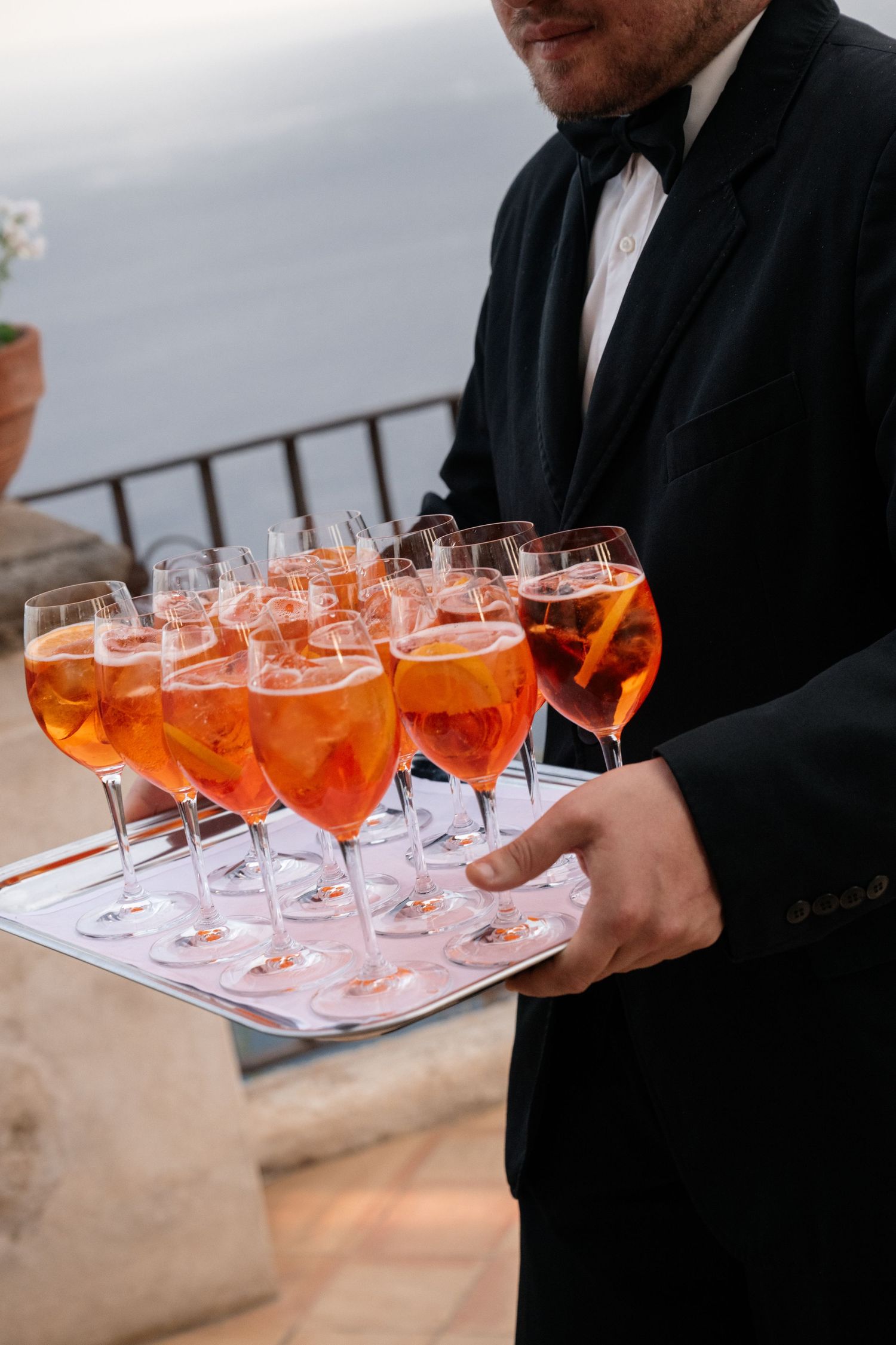 Waiter in black tuxedo serves tray of Aperol Spritz cocktails at elegant Italian wedding reception.