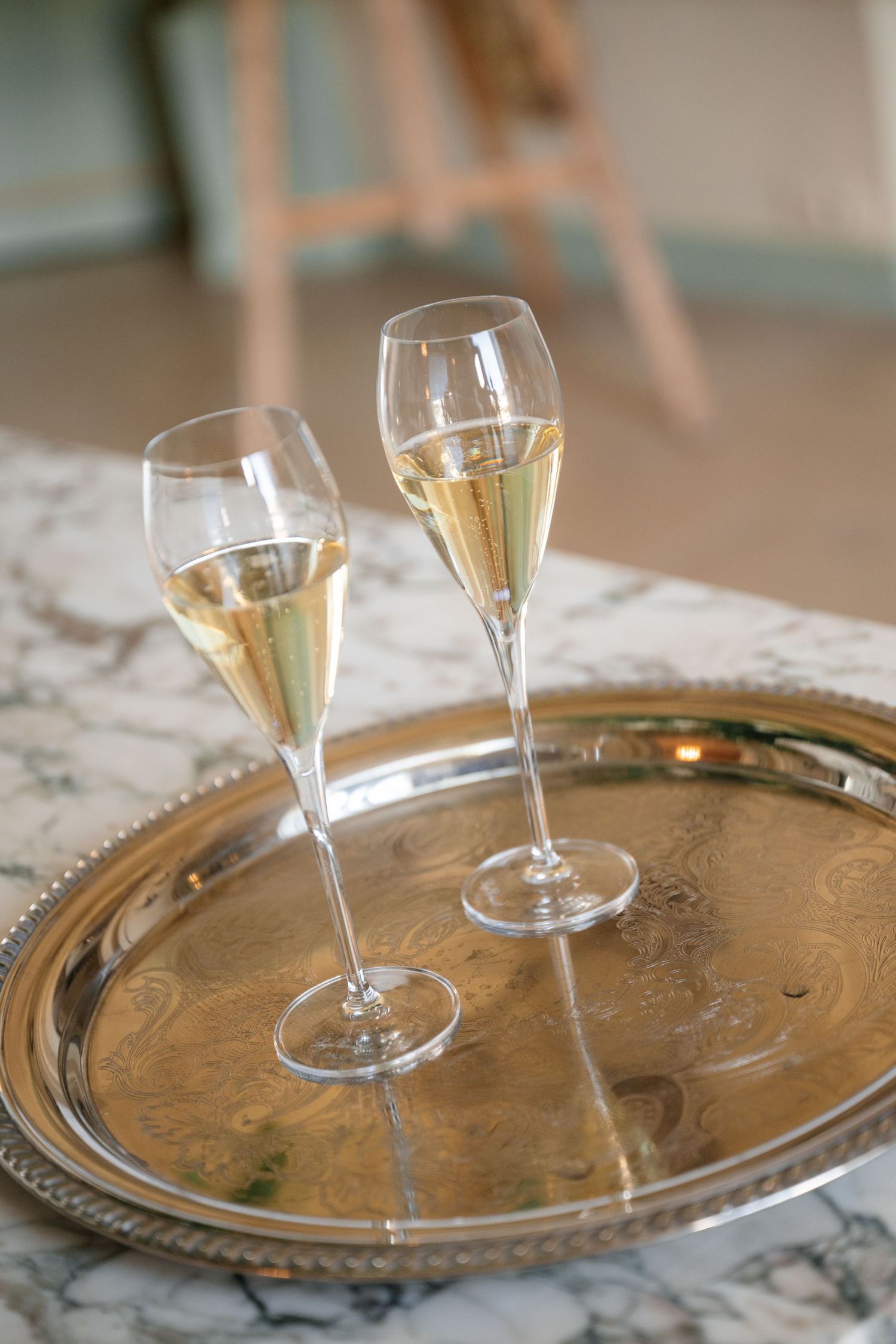 Two crystal champagne flutes filled with sparkling wine on an ornate silver tray on a marble surface.