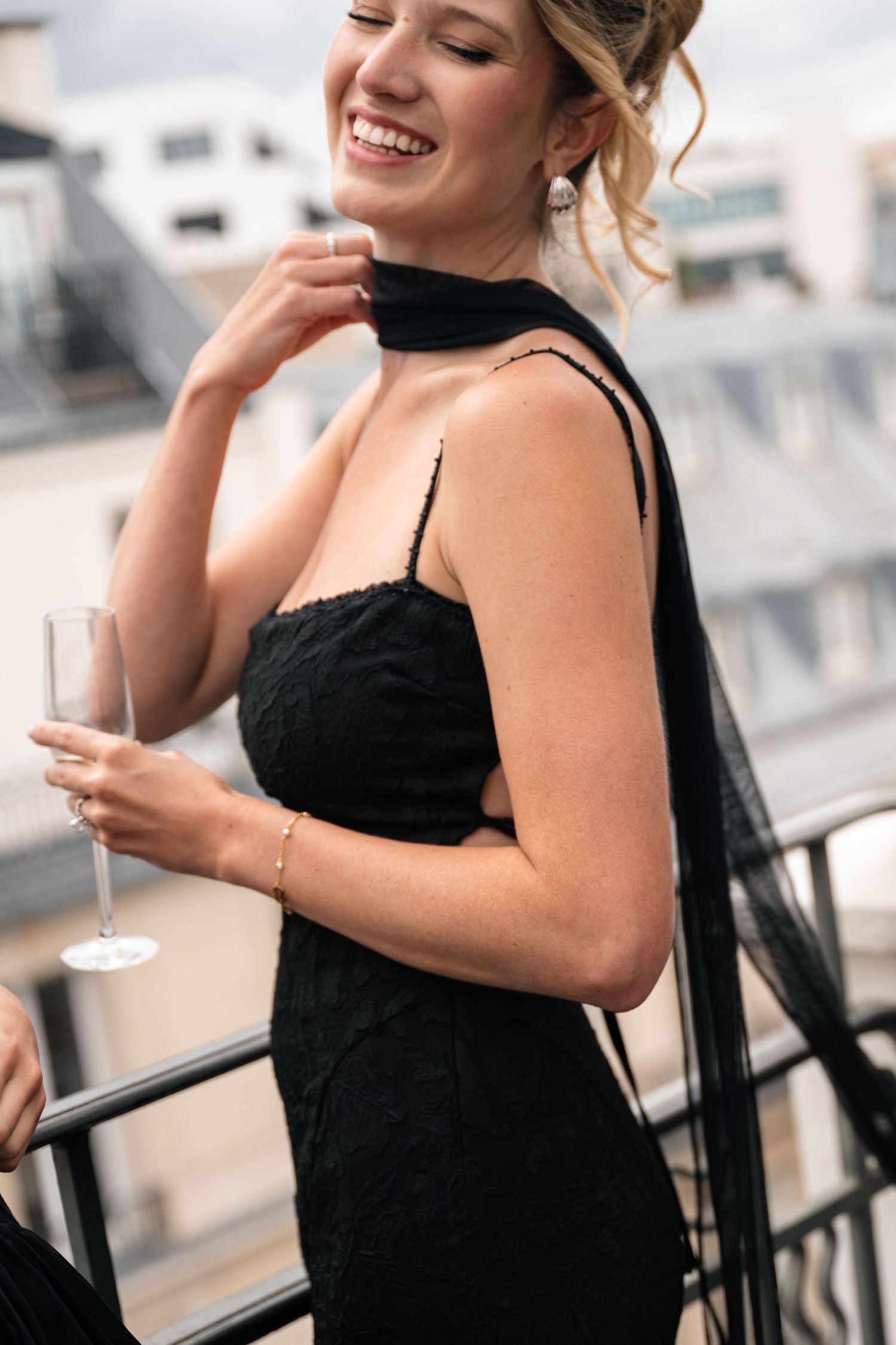 Elegant woman in a black lace dress smiling and holding a champagne flute on a rooftop terrace.