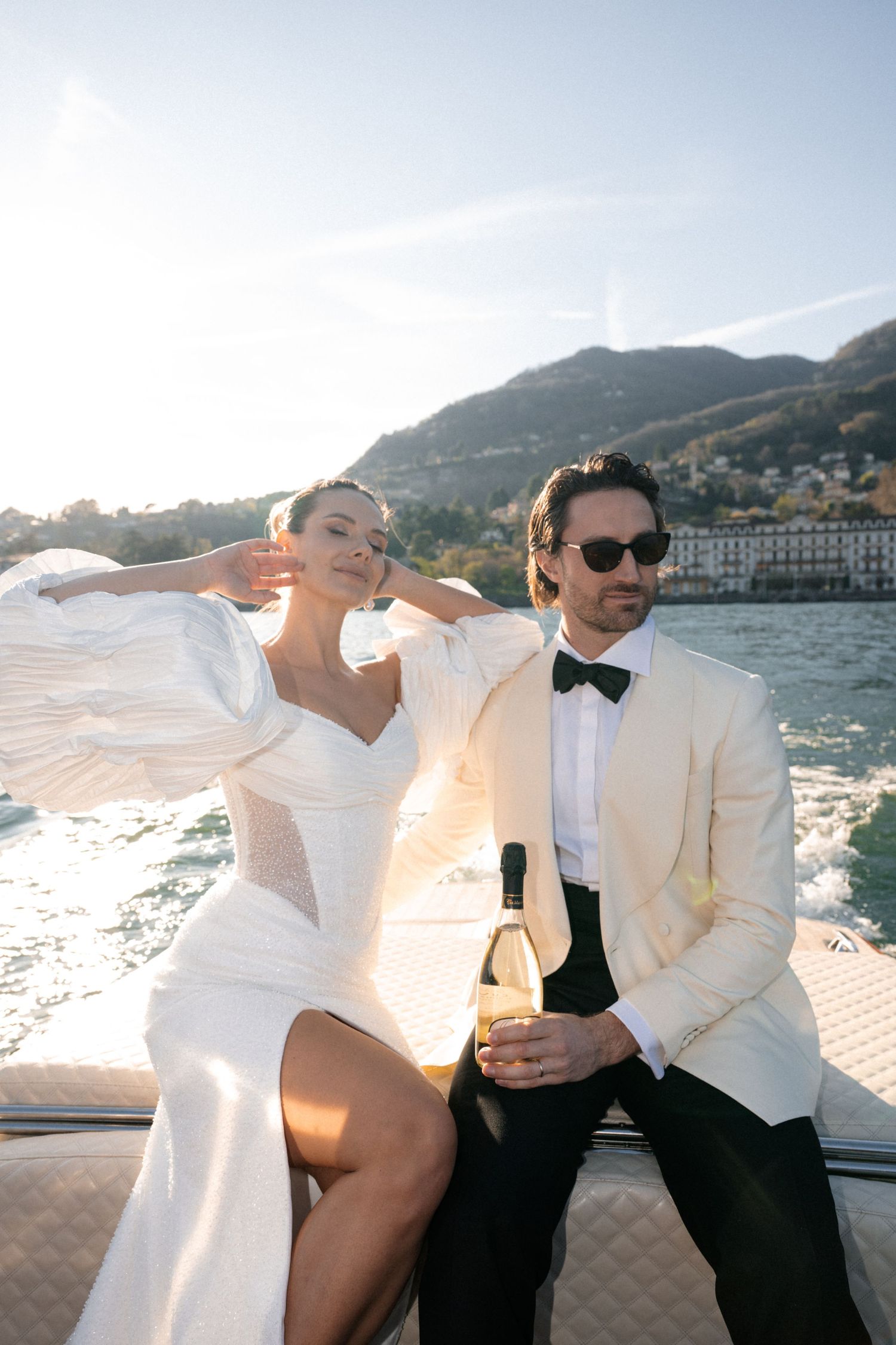 Elegant couple in wedding attire enjoy champagne on a speedboat against stunning Lake Como scenery.